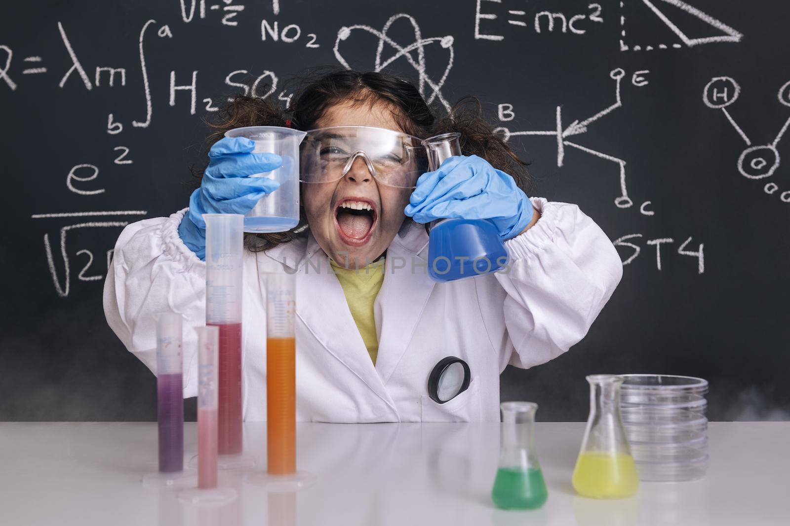 funny scientist girl with gloves and goggles in lab coat with chemical flasks, blackboard background with science formulas, explosion in the laboratory, back to school concept