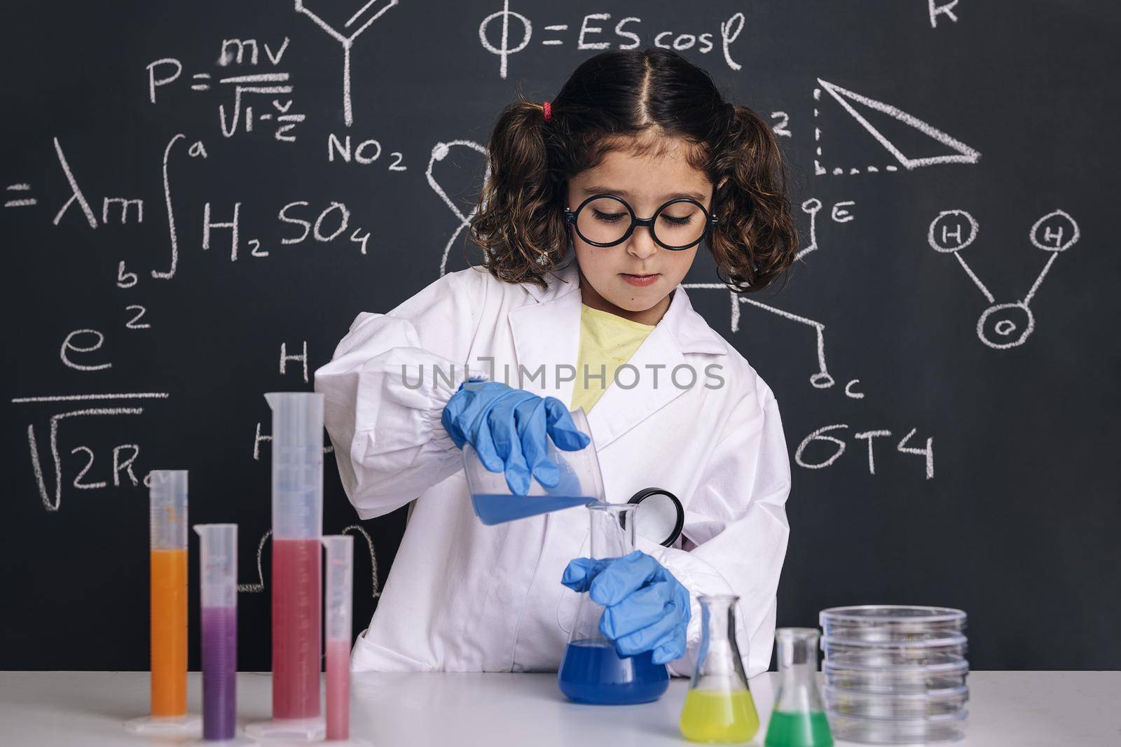 little scientist girl with glasses and gloves in lab coat mixing chemical liquids in flasks, on blackboard background with science formulas, concept of back to school and successful female career