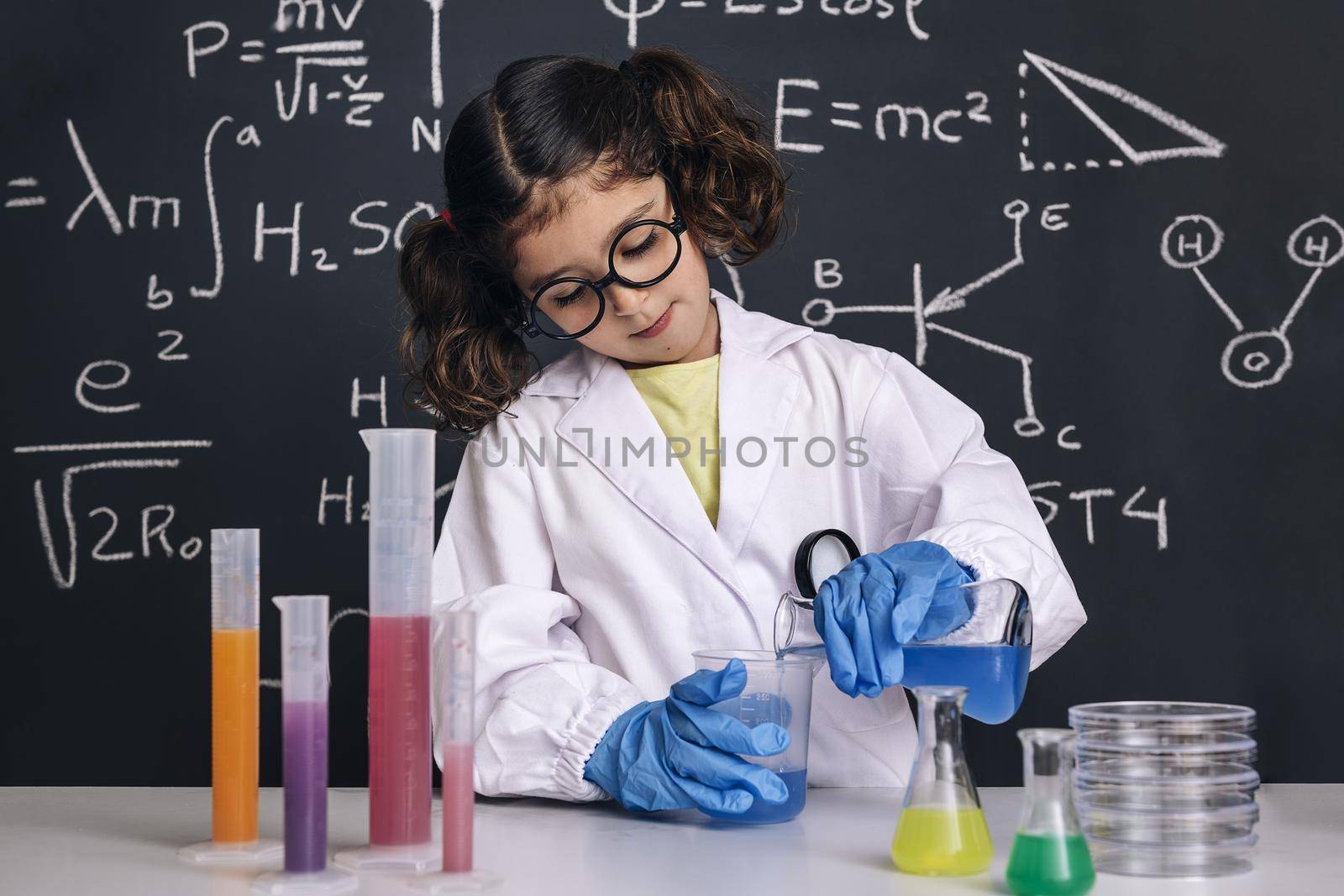 child scientist with glasses and gloves in lab coat mixing chemical liquids in flasks, on blackboard background with science formulas, concept of back to school and successful female career