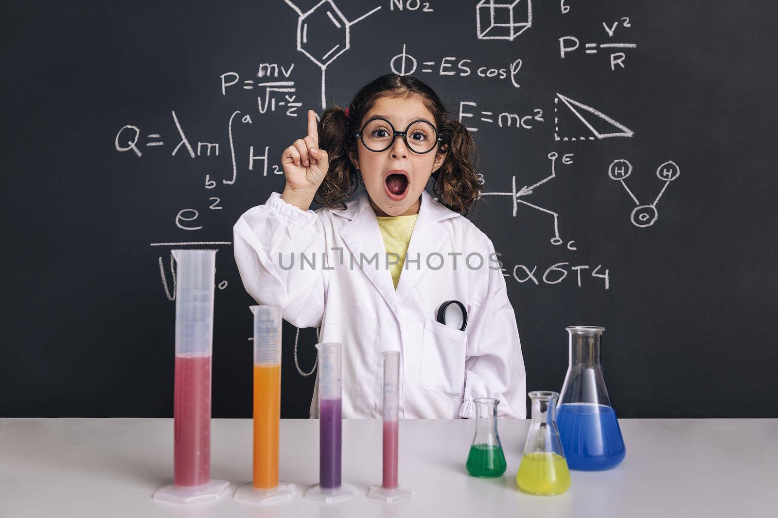 surprised scientist child with glasses in lab coat with chemical flasks having an idea, school blackboard background with science formulas, back to school and successful female career concept