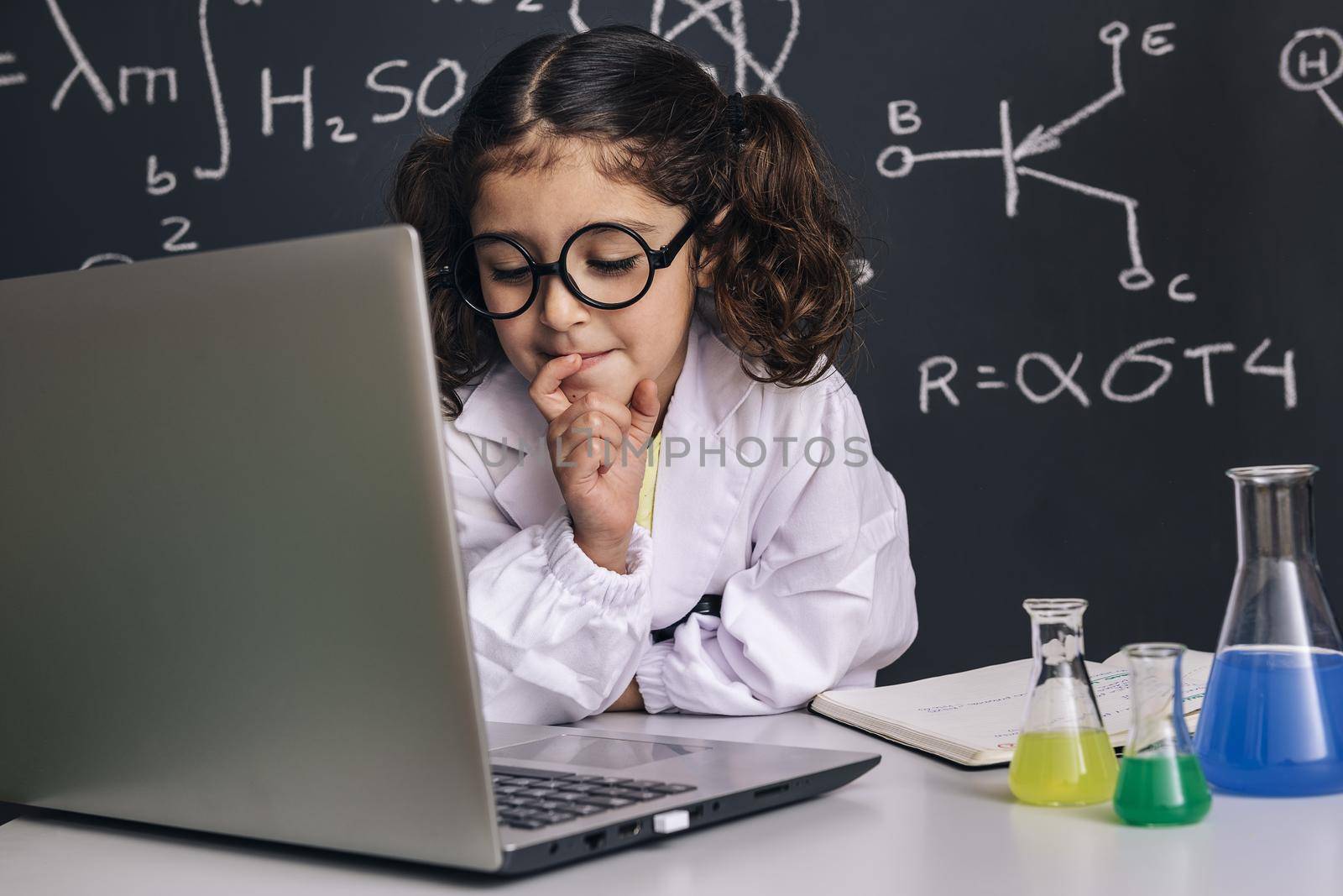 little scientist girl with glasses in lab coat looking to the computer, on school blackboard background with science formulas, back to school and successful female career concept