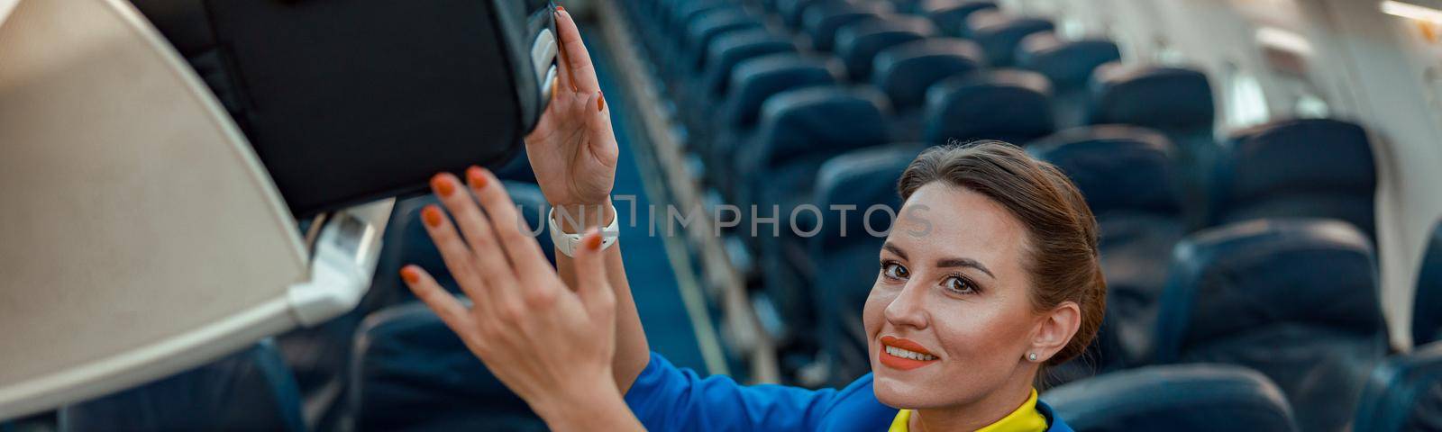 Smiling woman flight attendant placing travel suitcase in overhead baggage compartment while standing in aircraft passenger salon