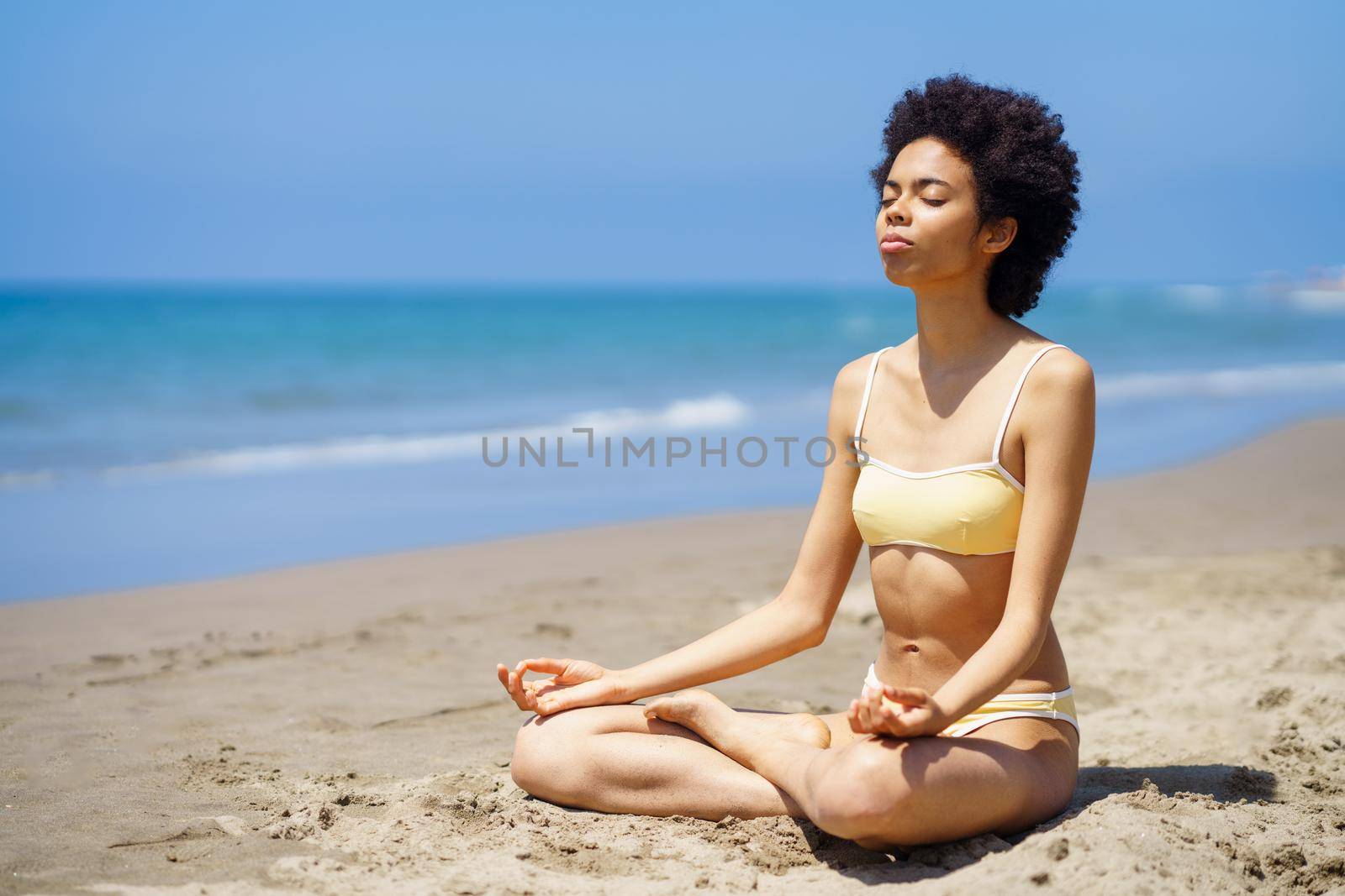 Full body of peaceful slim black female tourist in bikini sitting in Lotus pose on sandy seashore with closed eyes, while meditating during yoga session on sunny summer day