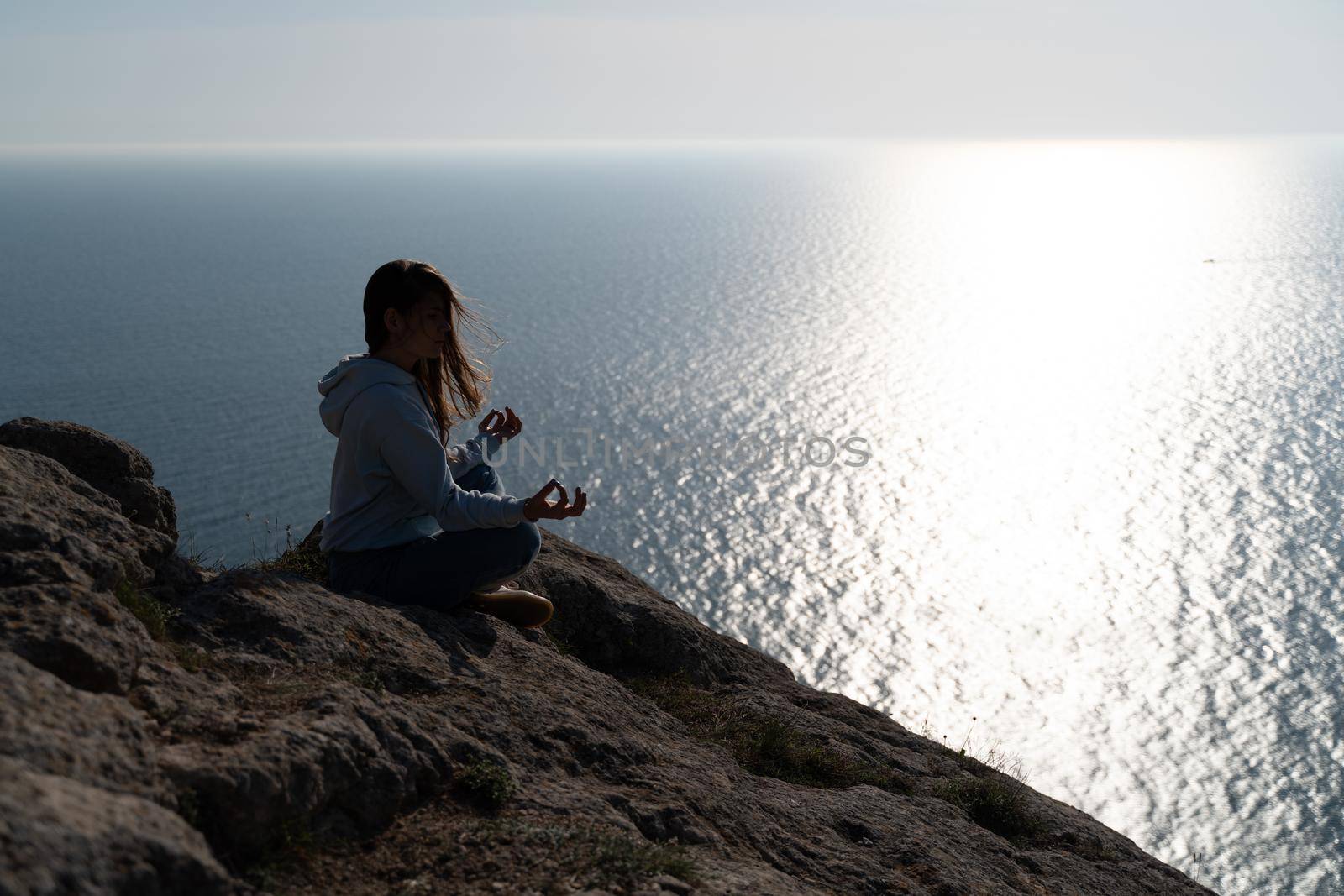 Woman tourist enjoying the sunset over the sea mountain landscape. Sits outdoors on a rock above the sea. She is wearing jeans and a blue hoodie. Healthy lifestyle, harmony and meditation.