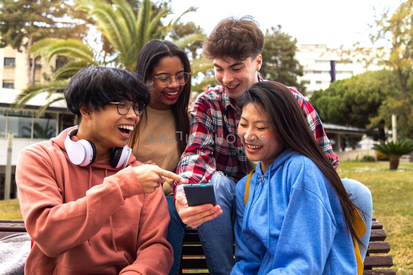 Happy young college student friends look at mobile phone. Multiracial teenagers using smartphone laughing together outdoor. Youth lifestyle and social media addiction concept.