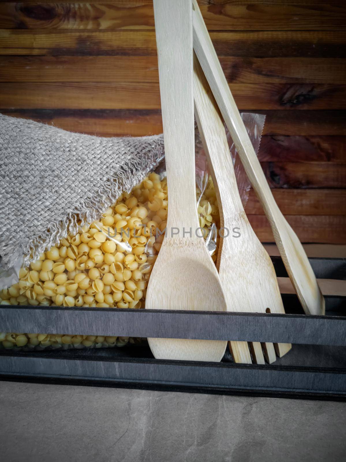 On the kitchen table, macaroni in transparent cellophane packaging, covered with a linen napkin, next to wooden Cutlery