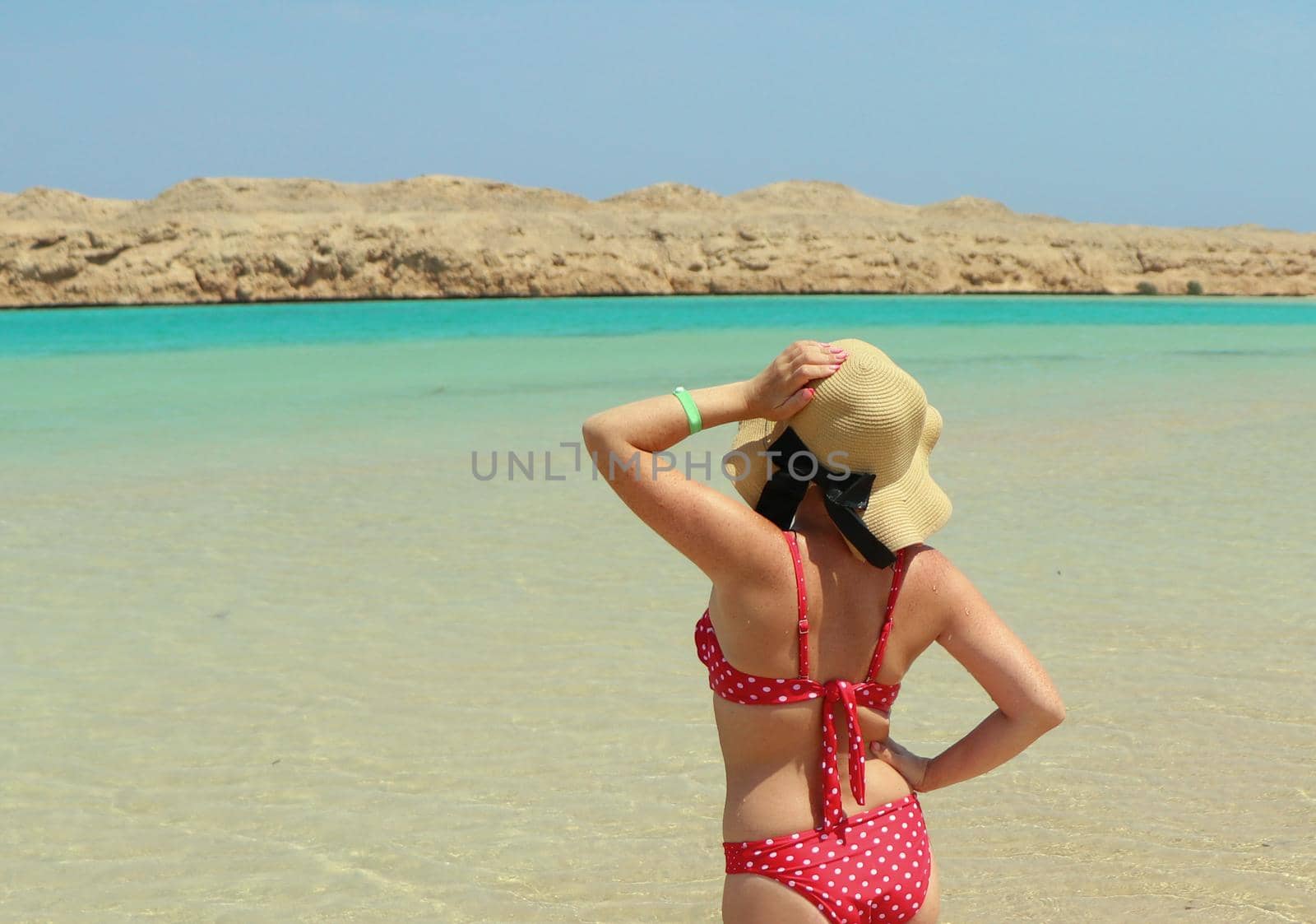 girl in a red swimsuit and a beach hat stands facing the sun in the sea. sunbathes standing in blue transparent water. Vacation concept by the sea, summer vacation. Blue sky . sunny summer day.