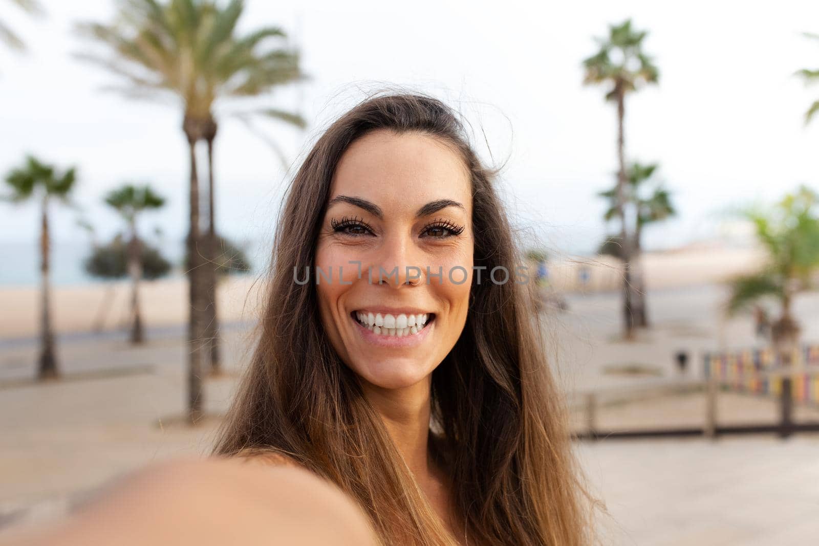 Young caucasian woman looking at camera taking selfie in the beach. Background with palm trees. Lifestyle and social media concept.