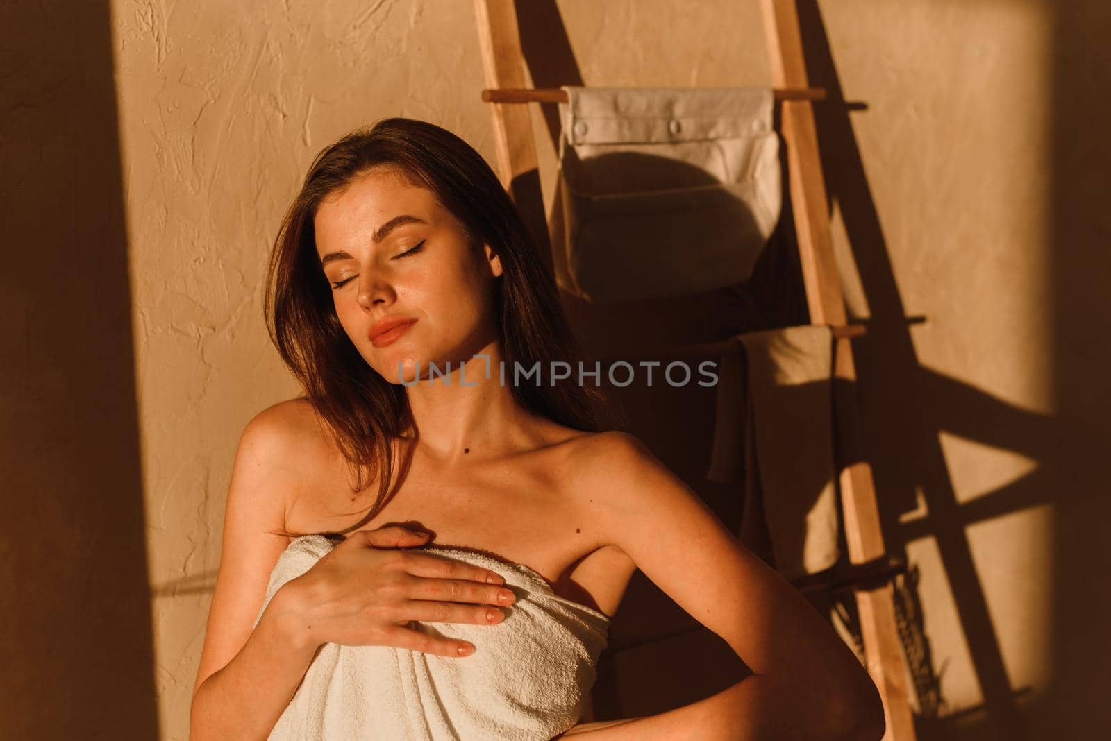 A young woman is enjoying the sun. A beautiful female model wrapped in a white towel stands in the bathroom after a shower. Shadows from tropical leaves