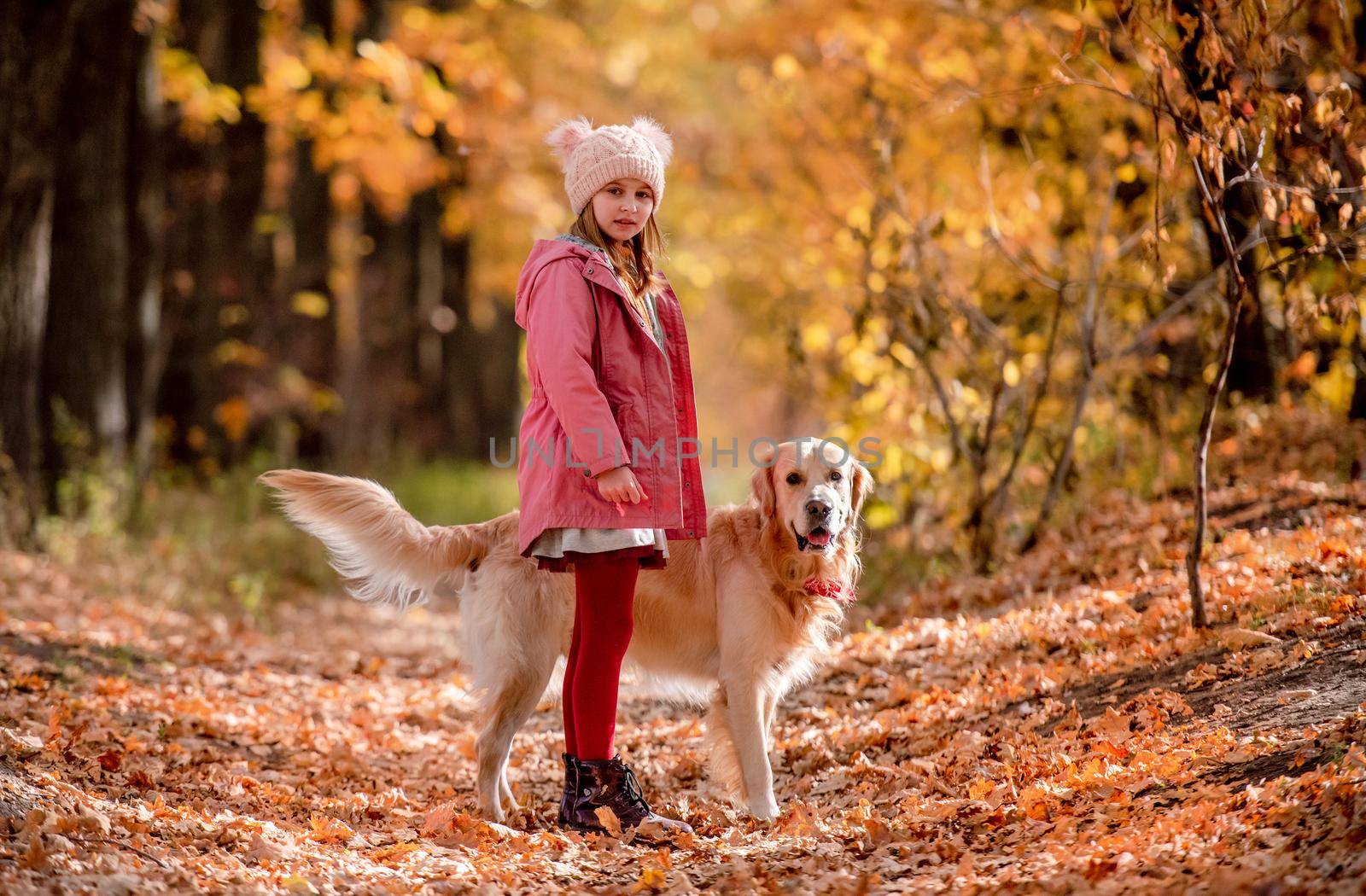 Cute girl kid with golden retriever dog looking at camera at autumn park. Beautiful portrait of child and pet outdoors at nature