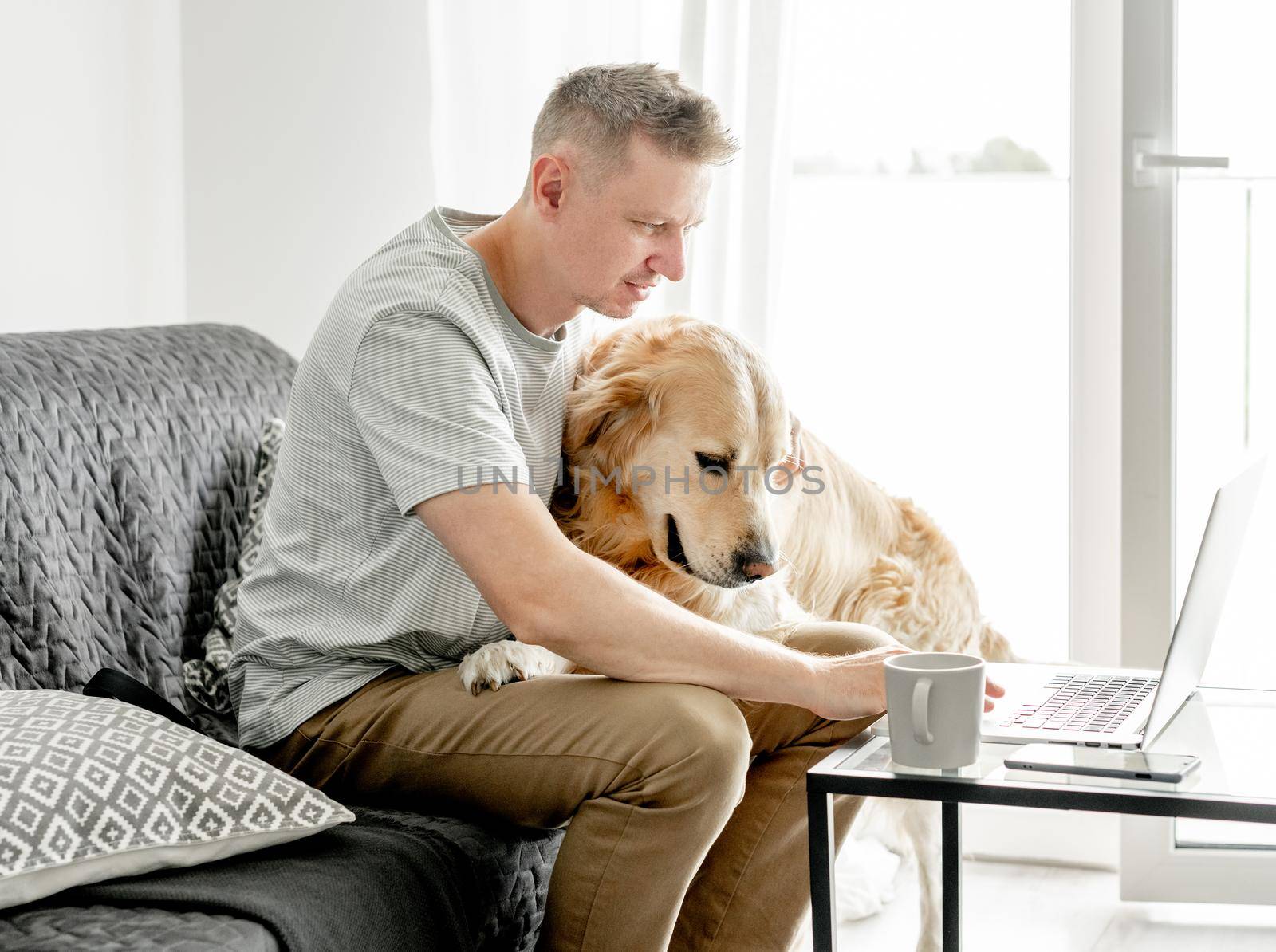 A young man works at a computer with his dog in a bright living room.