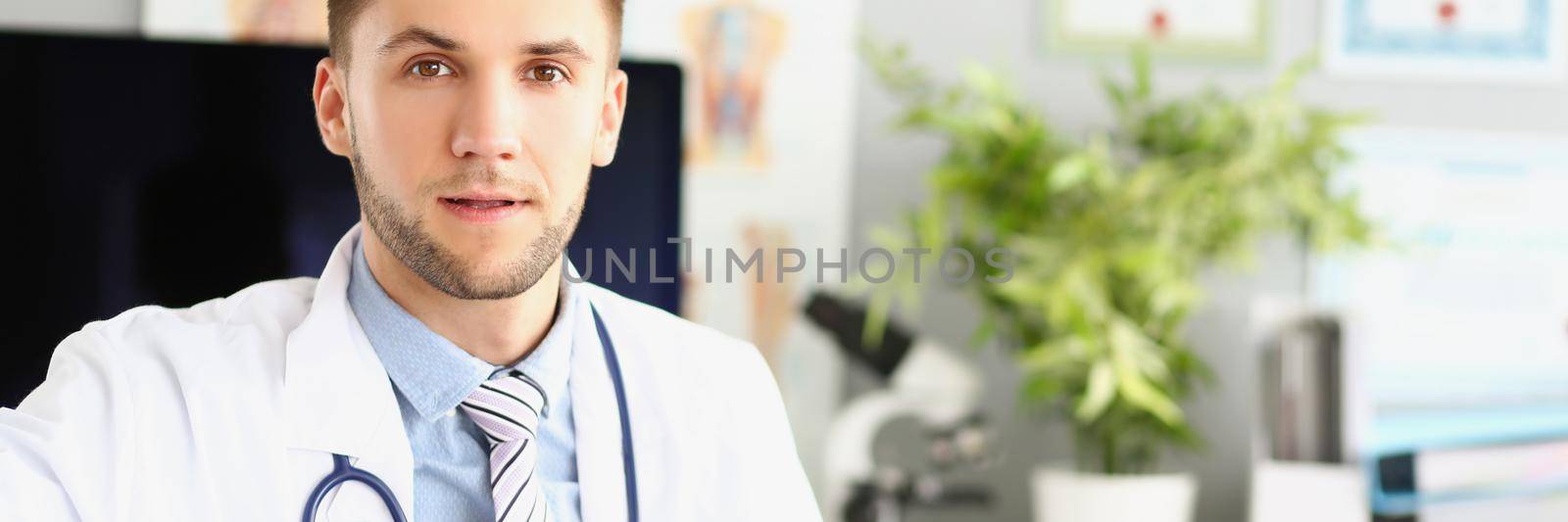 Handsome male doctor sitting at the table in the clinic, close-up, blurry background. Attentive doctor listens, portrait