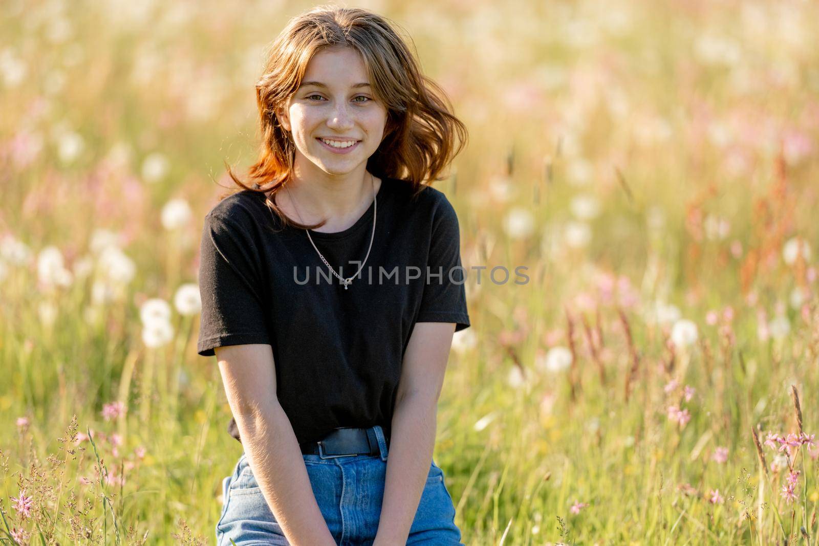 Pretty girl teenager sitting in field with dandelions and smiling outdoors. Beautiful young female person at nature in sunny day portrait