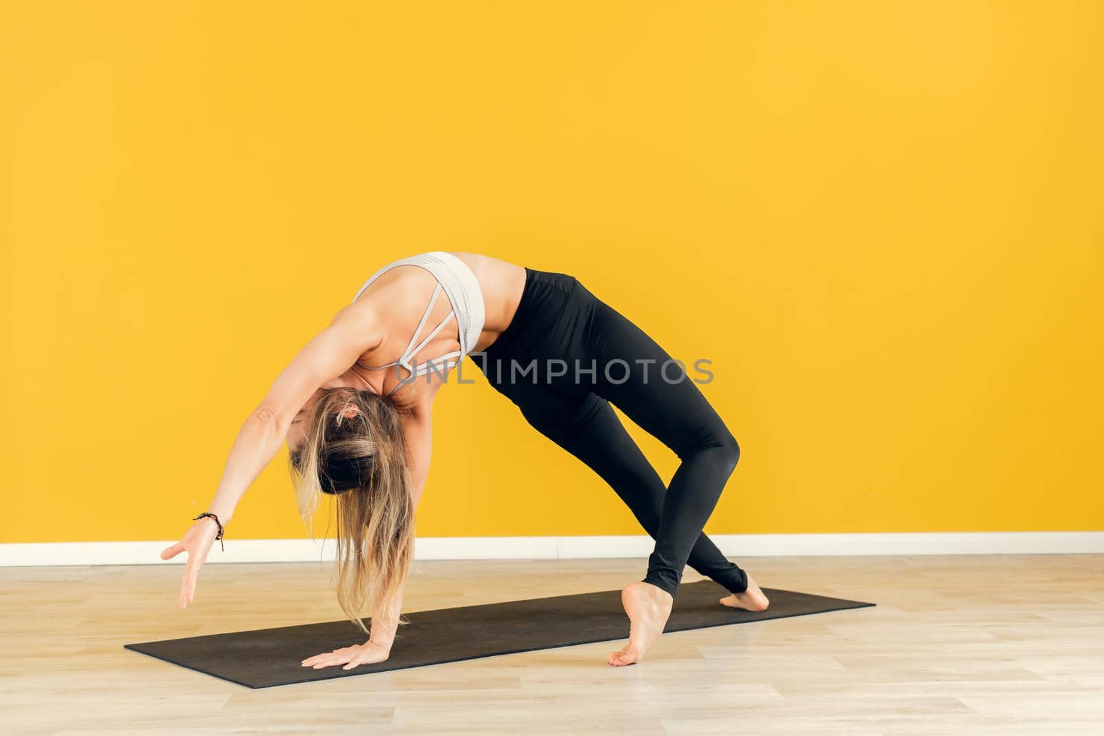 Young attractive woman practicing yoga, standing in one exercise, wearing black and white sportswear, yellow studio background, side view.