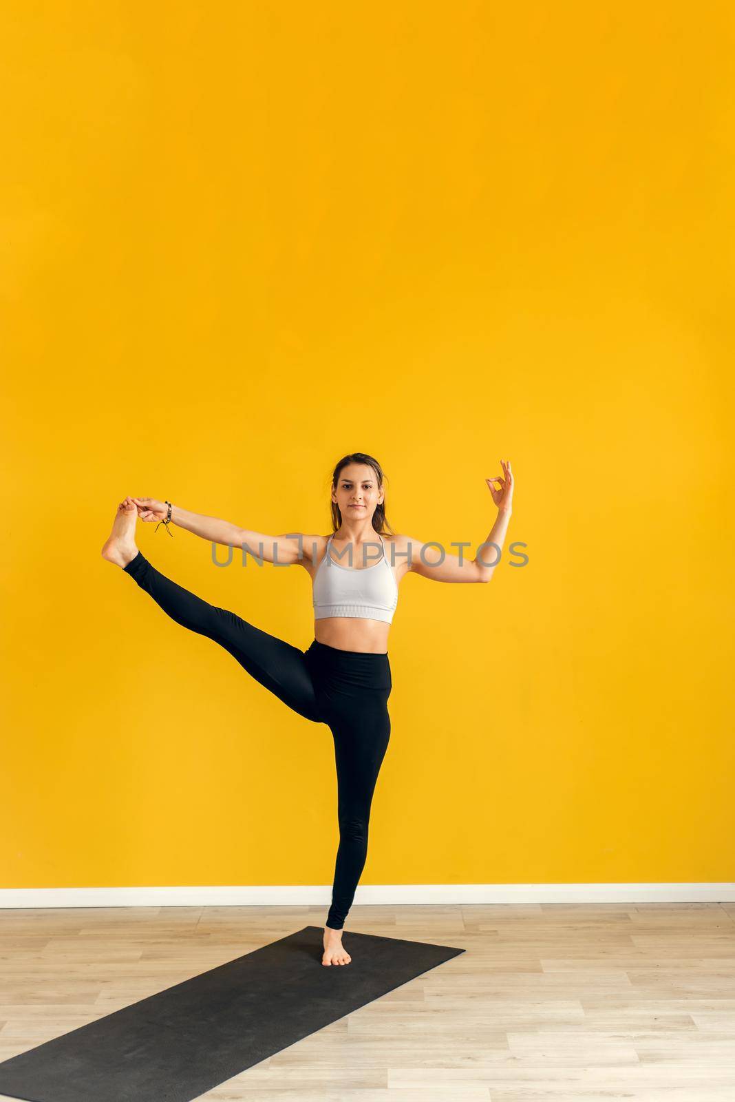 Beautiful young woman practicing yoga indoors. Utthita Hasta Padangusthasana, Extended Hand to Big Toe pose. Yellow background.