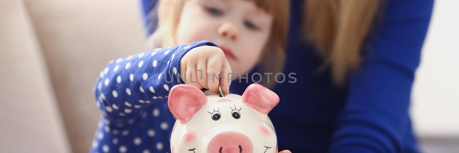 A little girl puts a coin in a pink piggy bank by kuprevich