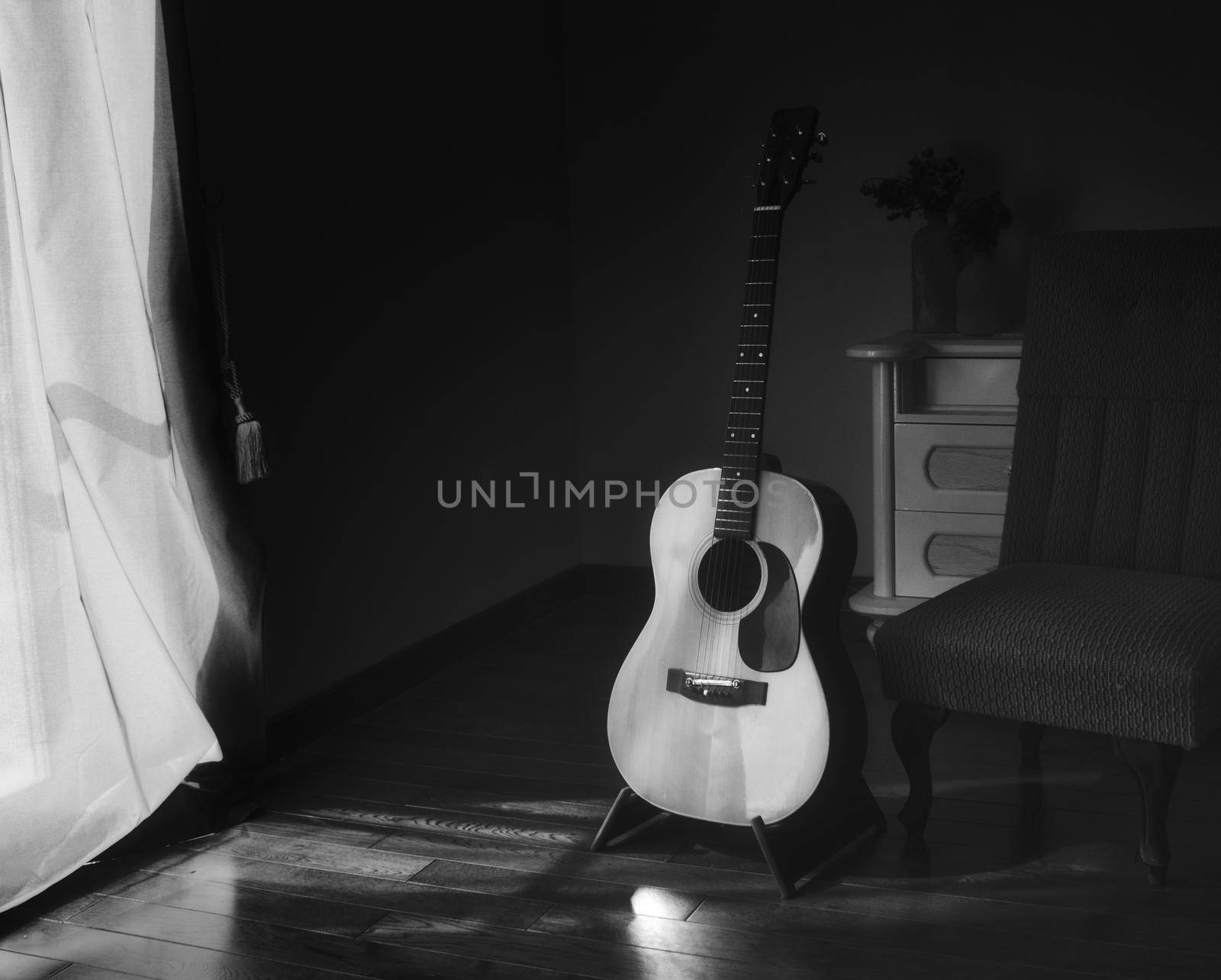 Black & white photo of acoustic Spanish guitar on a stand in the moody shadows of a dark room with bright light coming in from behind a curtain by tennesseewitney
