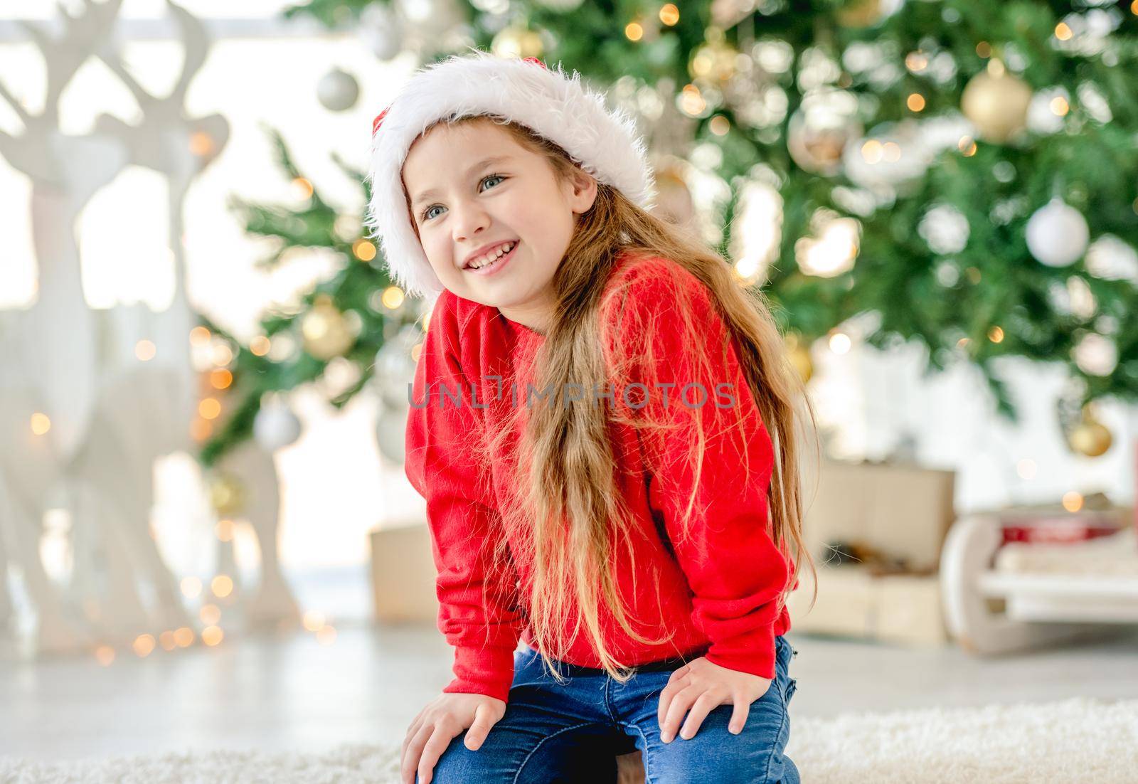 Little girl child wearing Santa hat in Christmas time sitting and smiling at decorated New Year home with festive tree. Cute kid in Xmas time