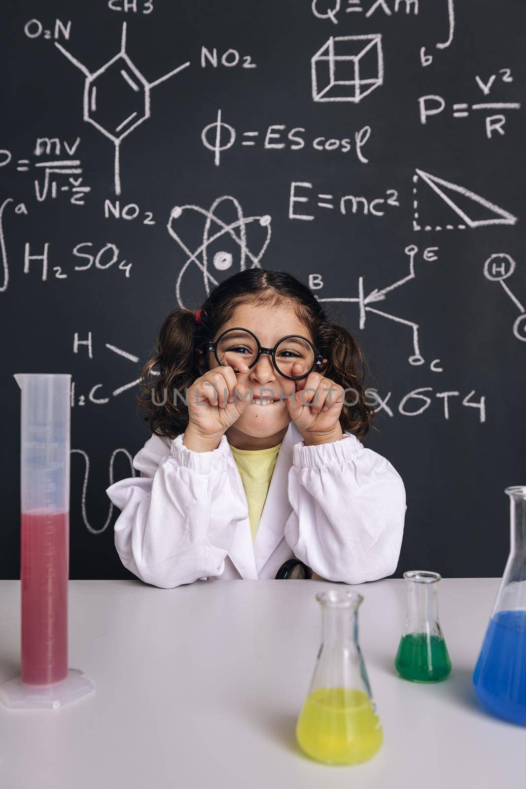 funny little scientist girl in lab coat joking with her glasses, on blackboard background with science formulas, concept of back to school and successful female career