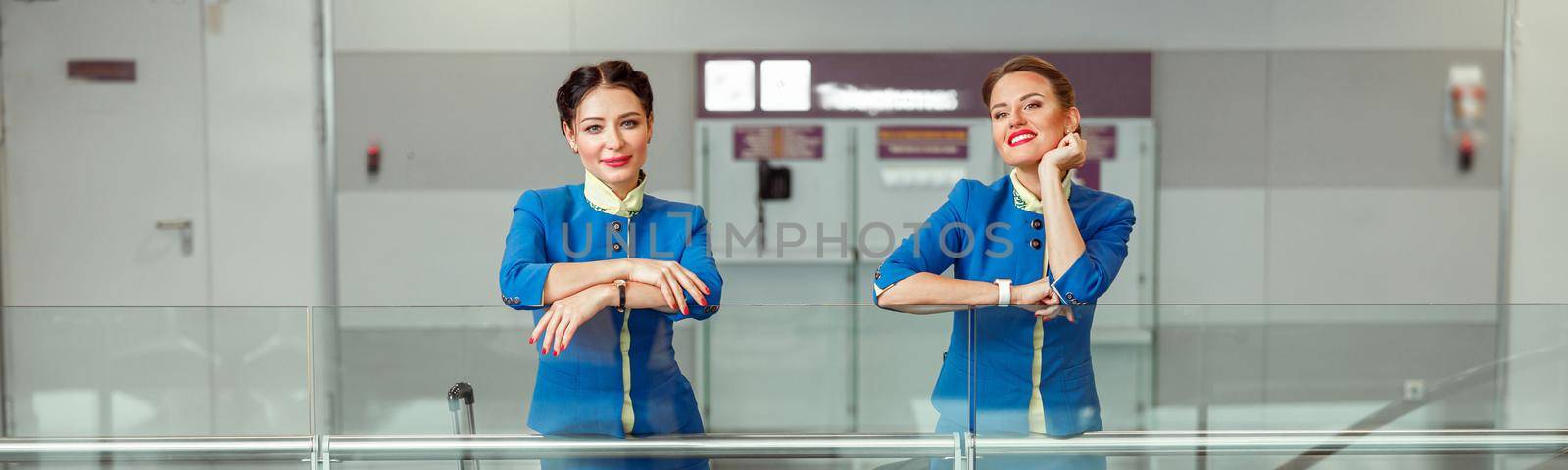 Cheerful female flight attendants in air hostess uniform looking at camera and smiling while standing near trolley luggage bags