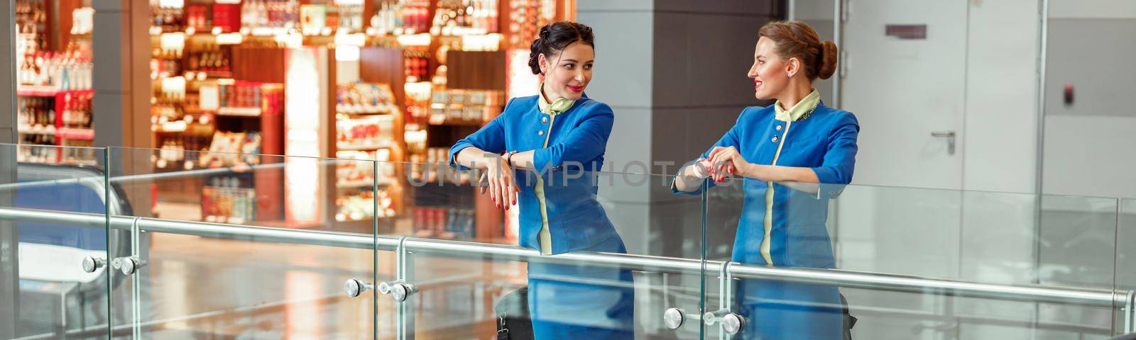 Women stewardesses in aviation air hostess uniform talking and smiling while standing near travel bags in passenger departure terminal