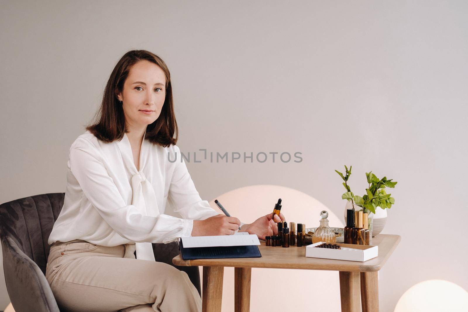 The aromatherapist girl is sitting in her office and holding a bottle of aromatic oil in her hands and writing something down. there are essential oils on the table.