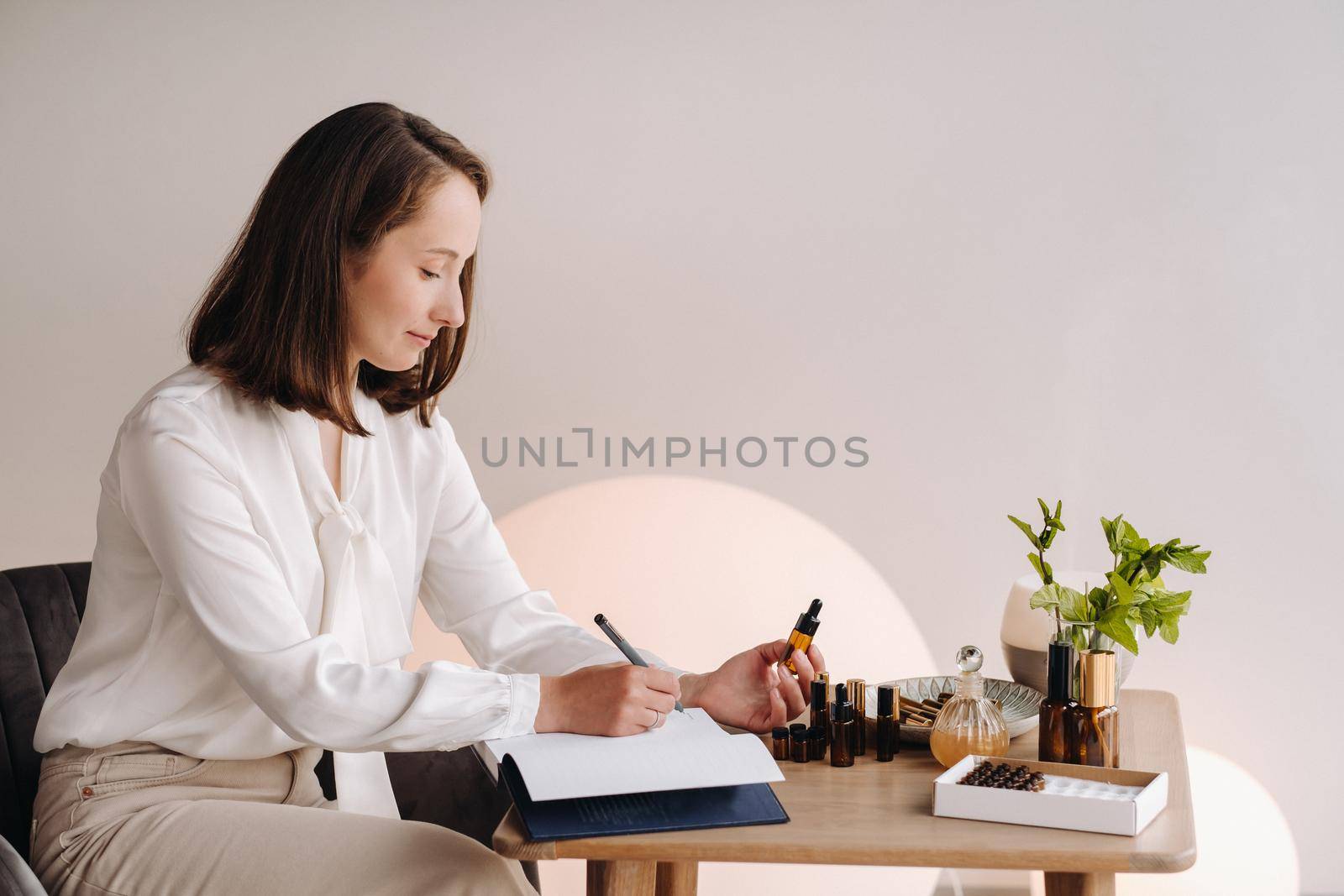 The aromatherapist girl is sitting in her office and holding a bottle of aromatic oil in her hands and writing something down. there are essential oils on the table by Lobachad