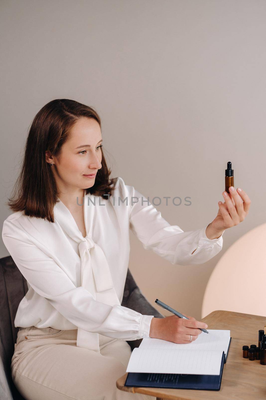 The aromatherapist girl is sitting in her office and holding a bottle of aromatic oil in her hands and writing something down. there are essential oils on the table.