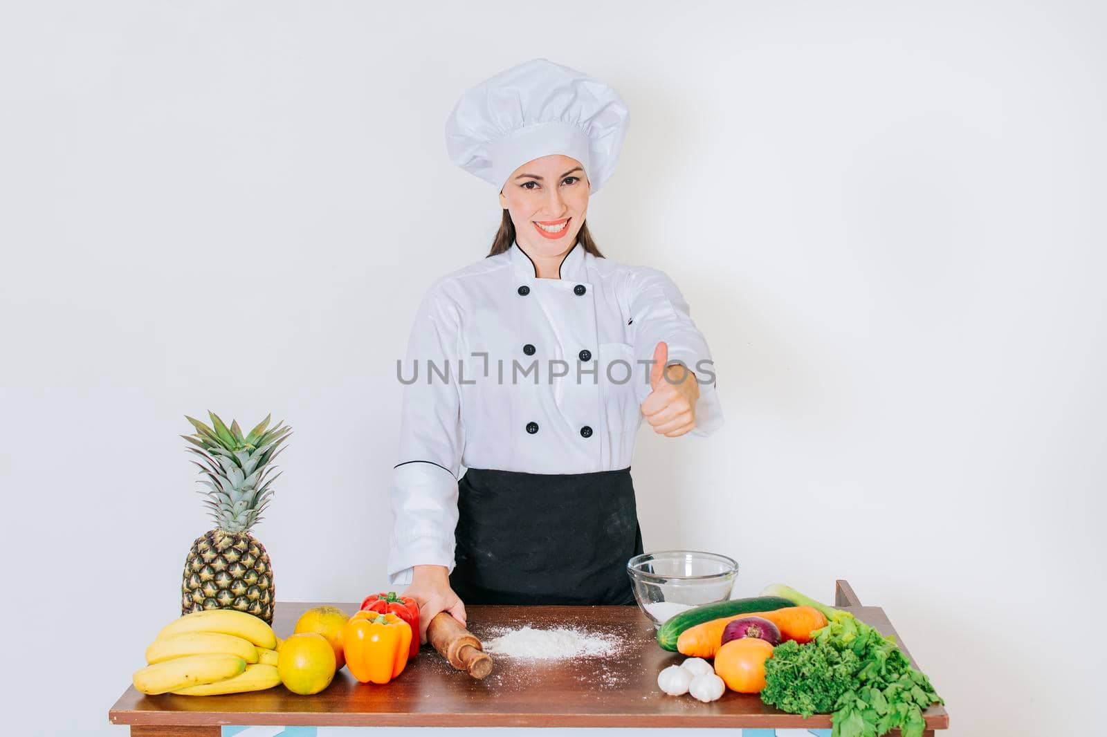 Chef woman holding bread rolling pin with thumb up, Smiling chef woman holding rolling pin on vegetable table, Concept of chef woman holding bread rolling pin by isaiphoto