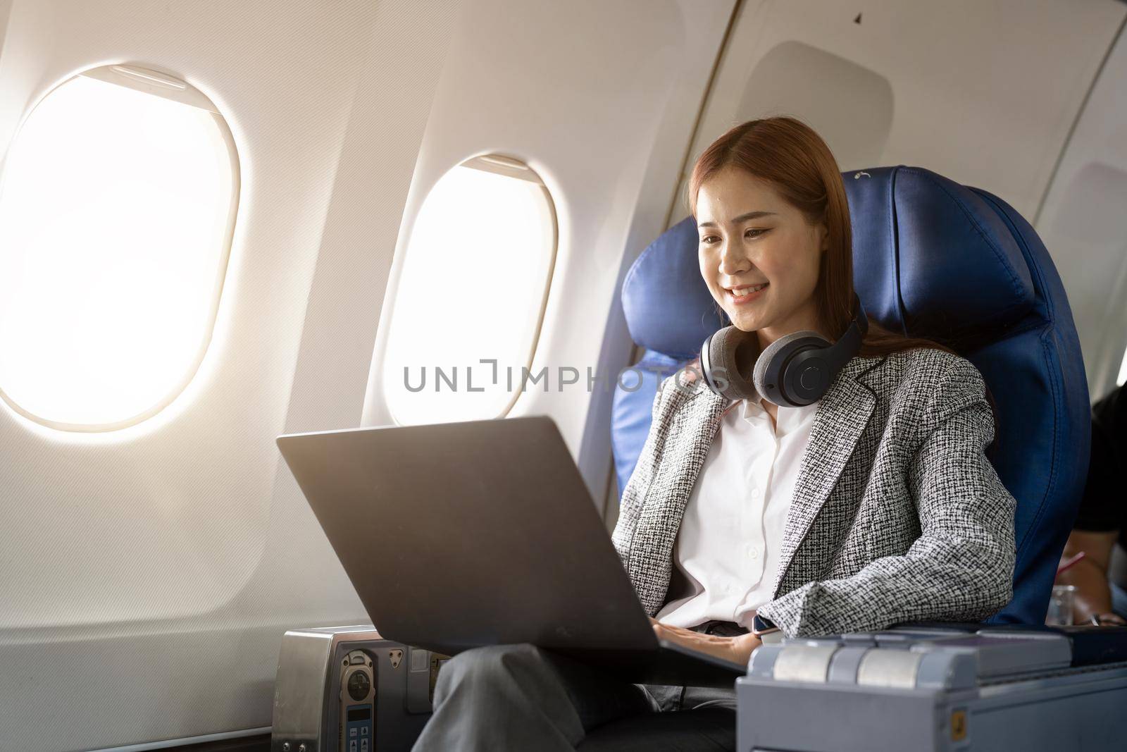 Asian young woman wearing headphone using laptop sitting near windows at first class on airplane during flight, Traveling and Business concept.