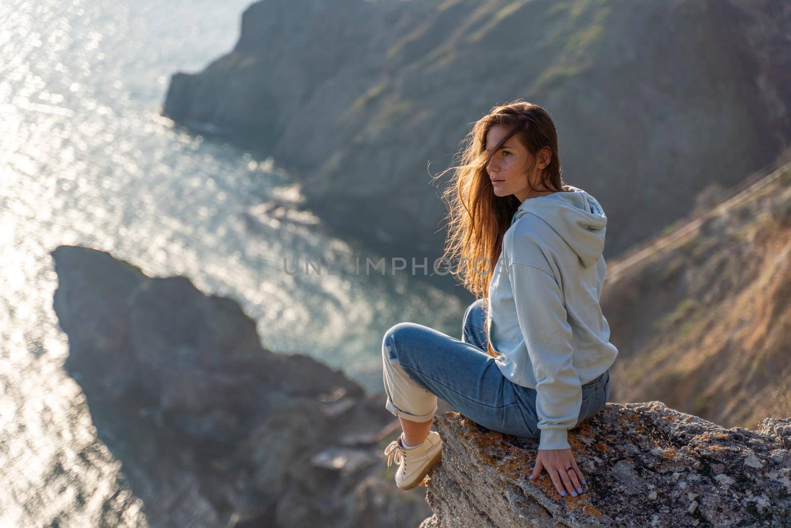 Woman tourist enjoying the sunset over the sea mountain landscape. Sits outdoors on a rock above the sea. She is wearing jeans and a blue hoodie. Healthy lifestyle, harmony and meditation by Matiunina