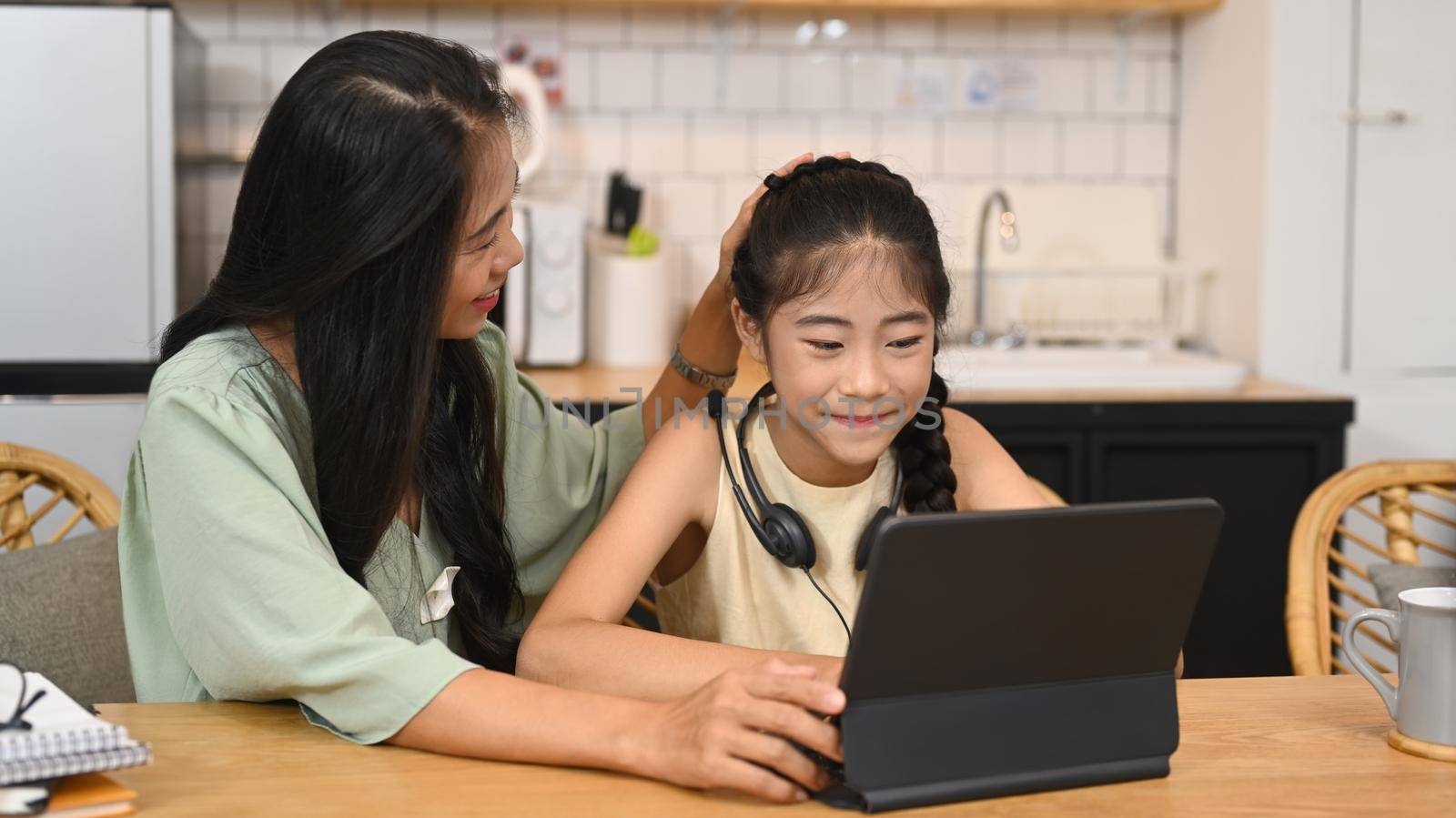 Adorable little asian girl doing homework, learning online at home and her mother sitting nearby and giving support.