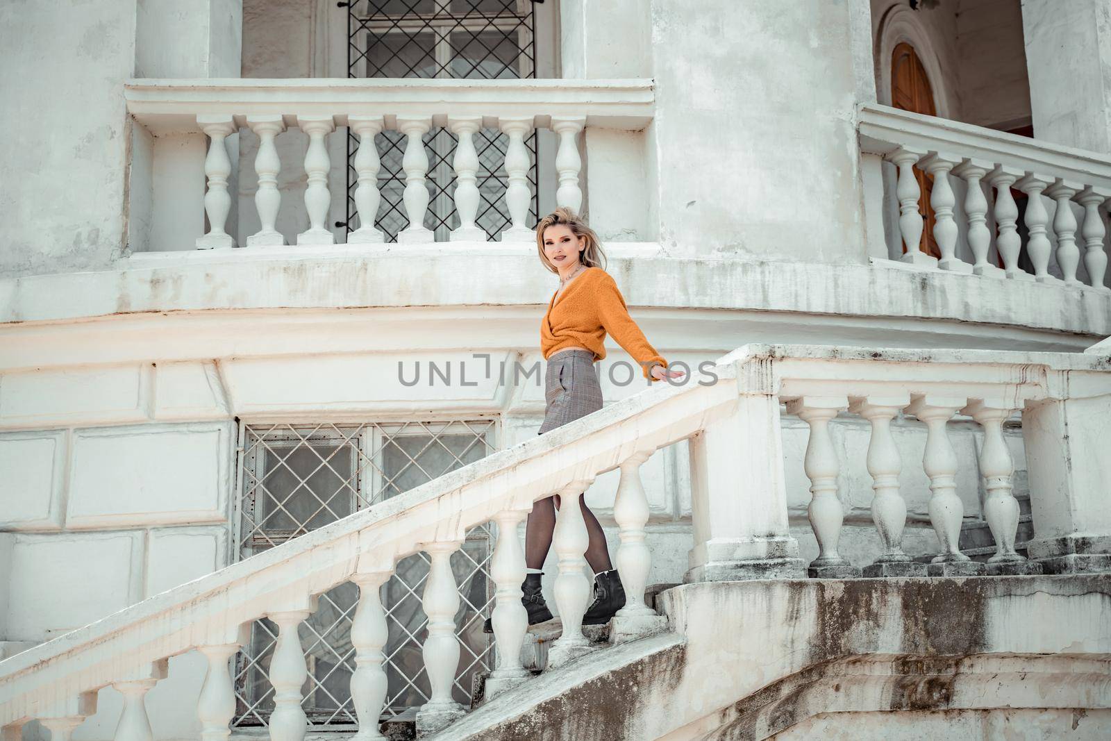 A middle-aged woman looks like a good blonde with curly beautiful hair and makeup on the background of the building. She is wearing a yellow sweater