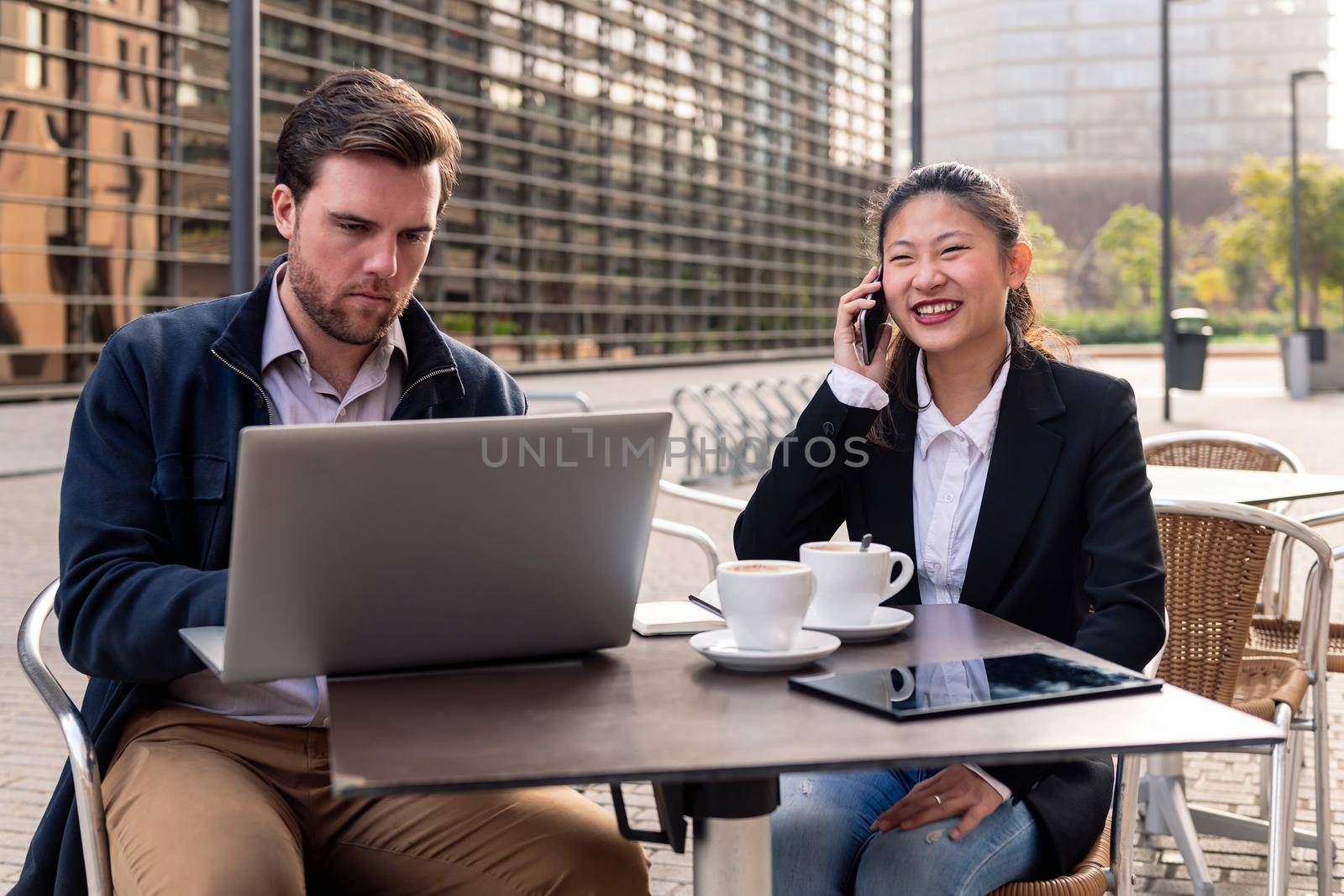 smiling asian businesswoman talking by phone over coffee and his partner work with the laptop on a terrace in the financial district, concept of entrepreneurship and business