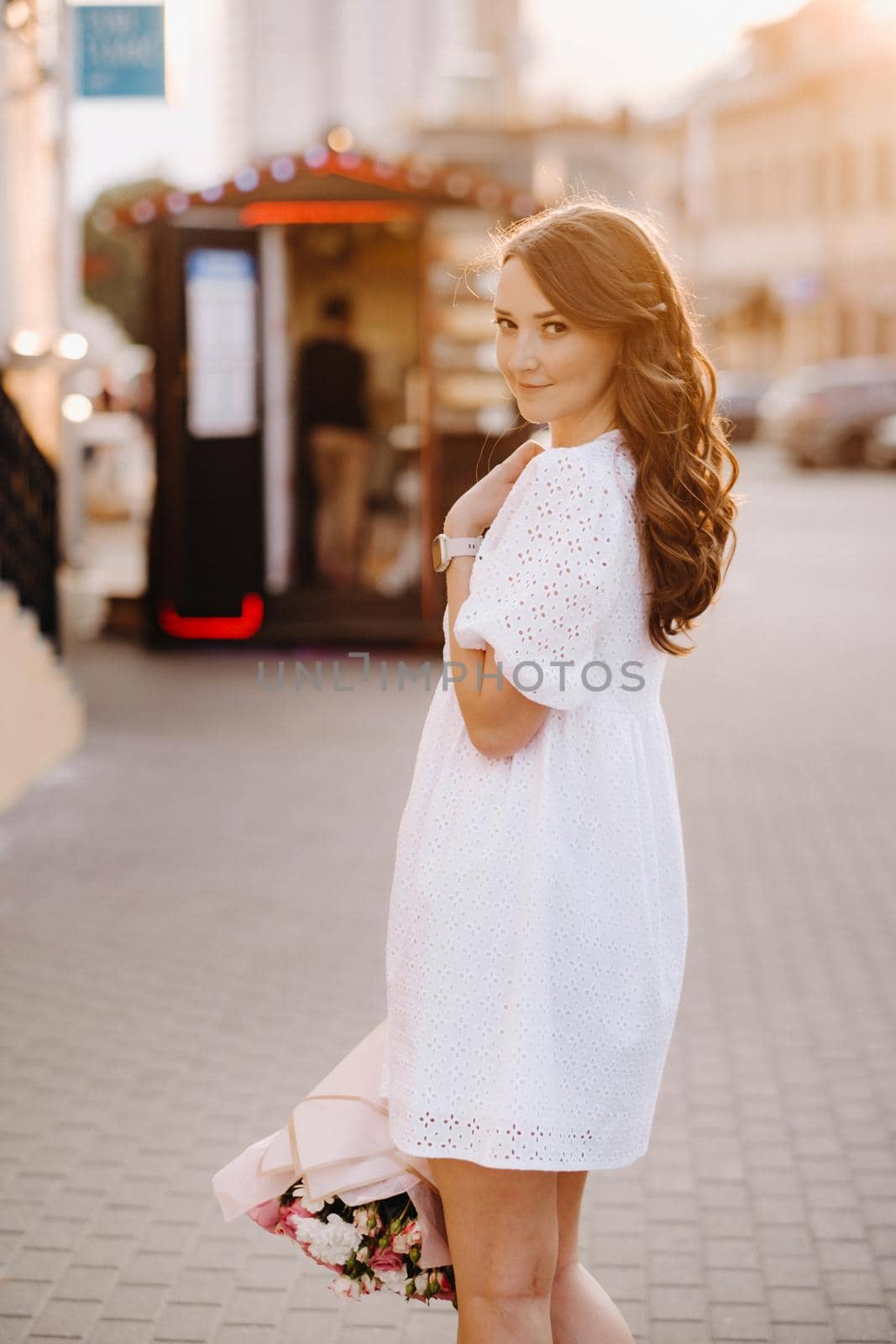 A happy woman in a white dress at sunset with a bouquet of flowers in the city.