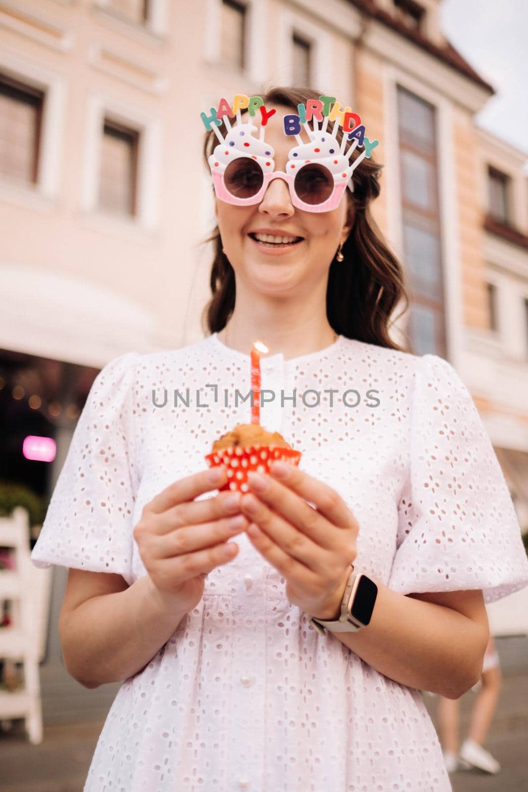 A beautiful happy woman in a white dress holds a cake in her hands on the street of the city celebrating her birthday.