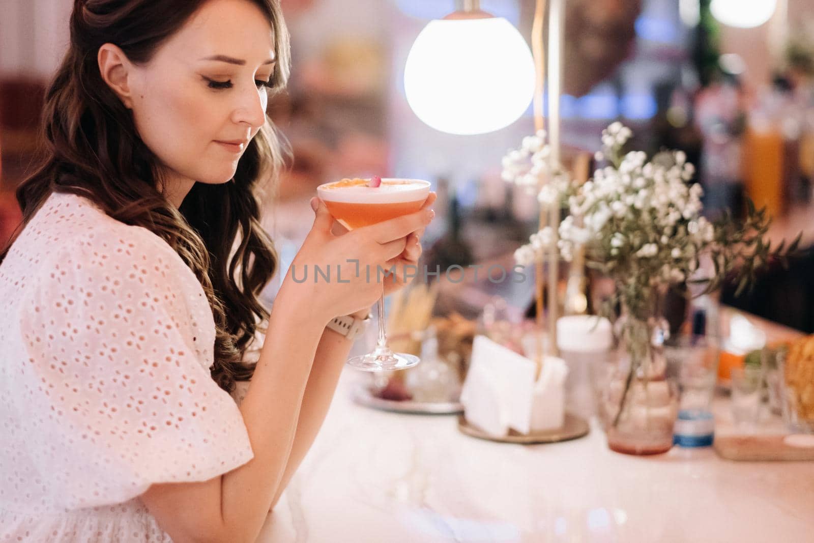 A girl in a white dress is sitting at the bar in a cafe and drinking a cocktail.