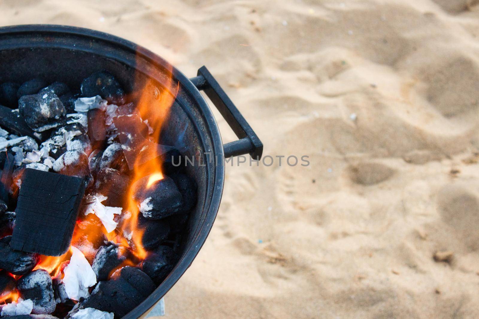 Closeup on charcoal burning with a yellow flame in a round metallic barbeque on a sandy beach