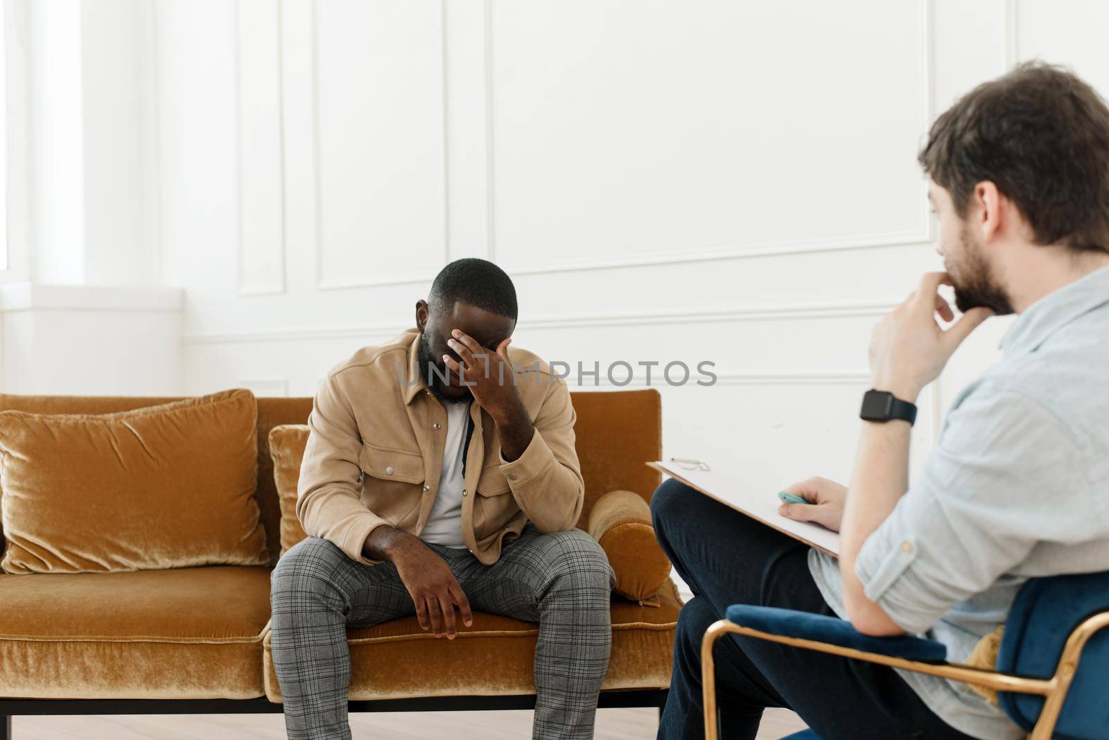 Frustrated young african american man sitting on sofa and holding hands near face in office at session with psychologist or psychiatrist. Desperate black man suffering from nervous breakdown or depression, having session at psychologist.