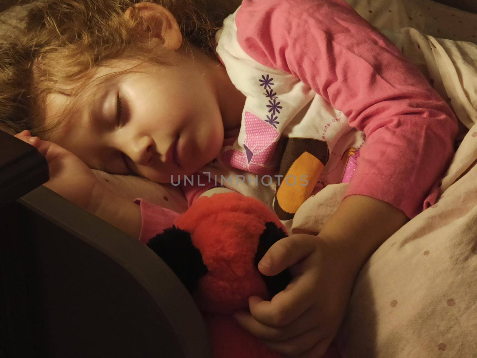 Adorable little girl asleep in her bed dimly lit by a bedside table lamp with a warm bulb by tennesseewitney