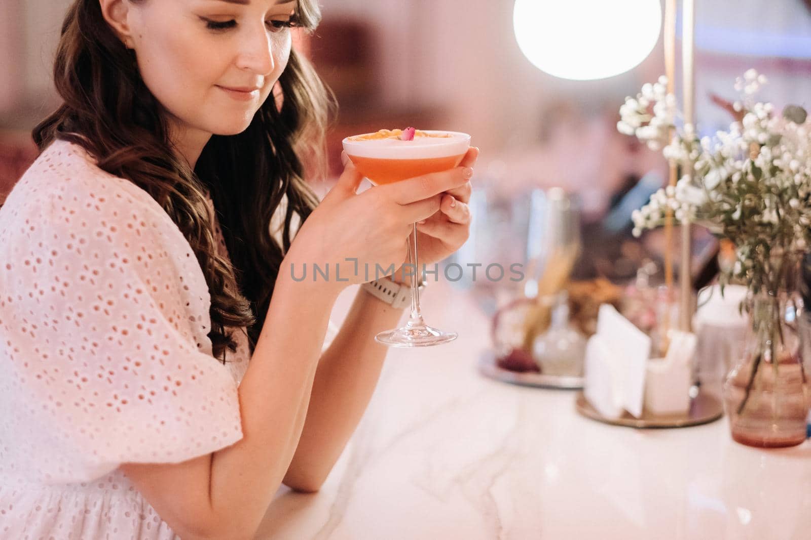 A girl in a white dress is sitting at the bar in a cafe and drinking a cocktail by Lobachad
