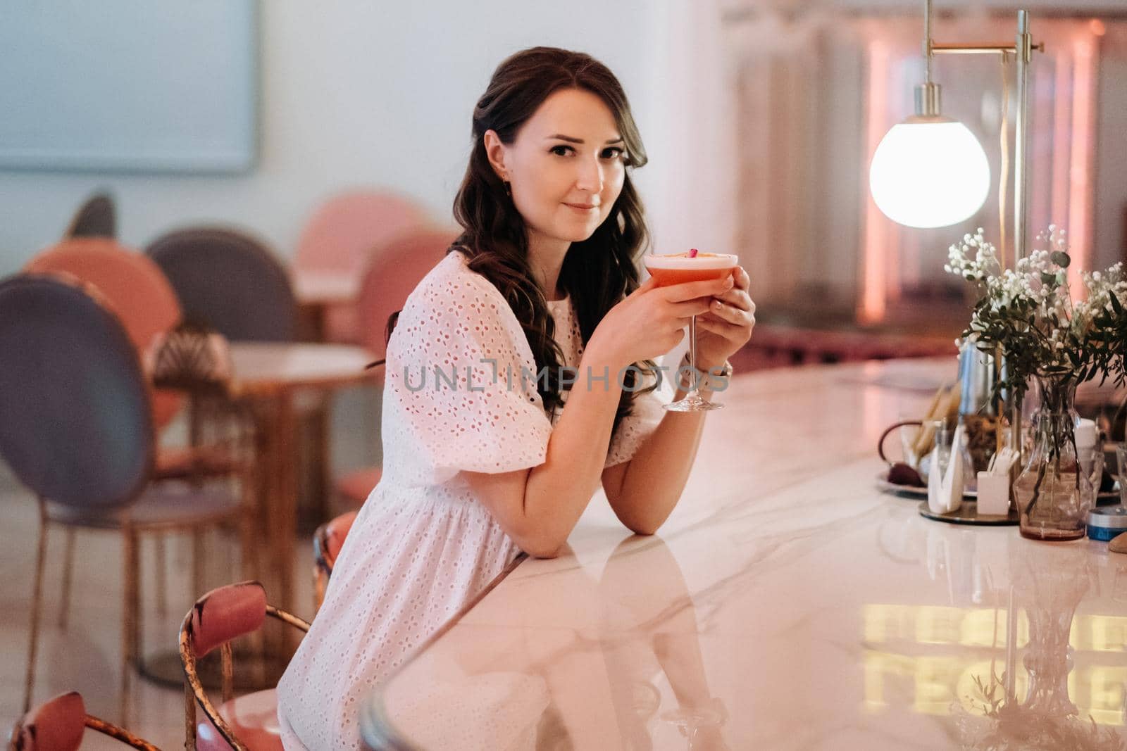A girl in a white dress is sitting at the bar in a cafe and drinking a cocktail by Lobachad