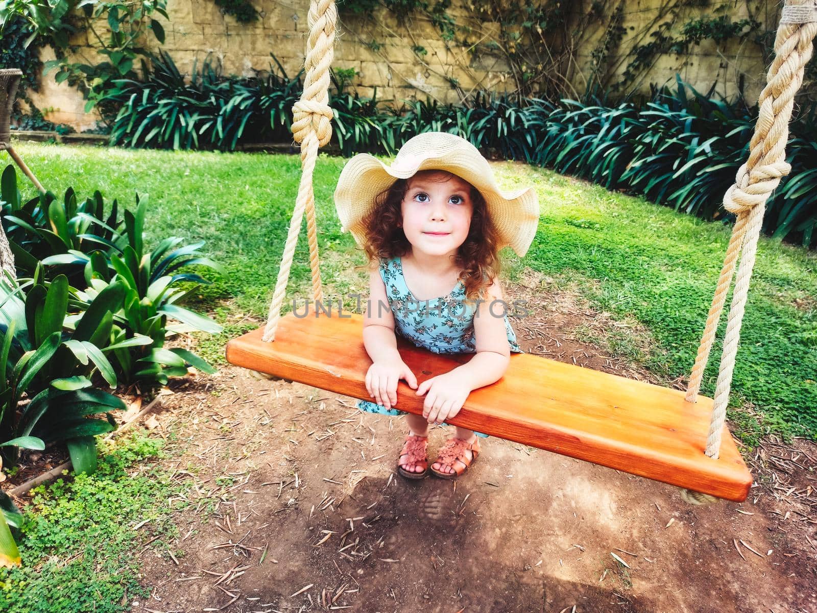 Cute little girl playing on a wooden swing wearing a big summer hat