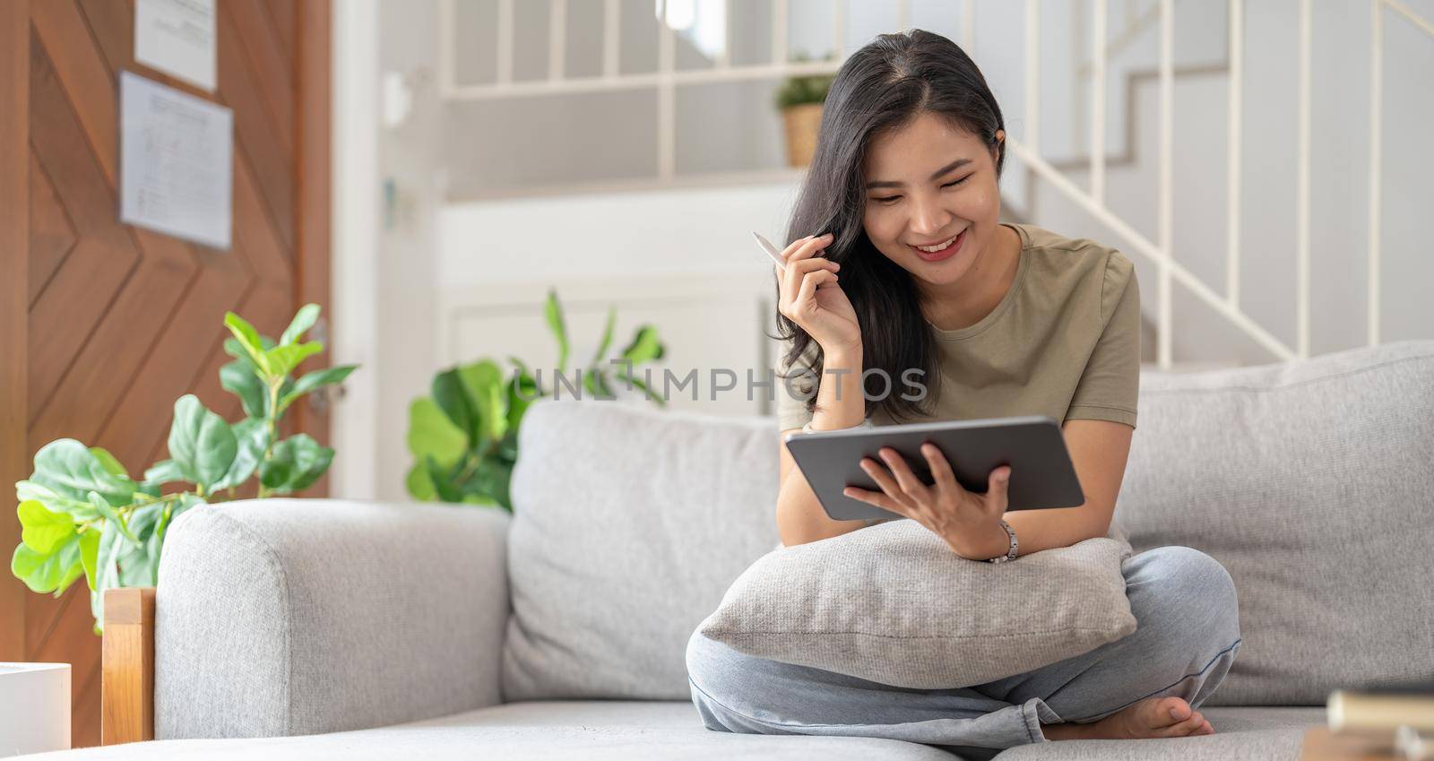 Beautiful attractive young Asian woman sitting on gray sofa and using digital tablet at living room in the morning