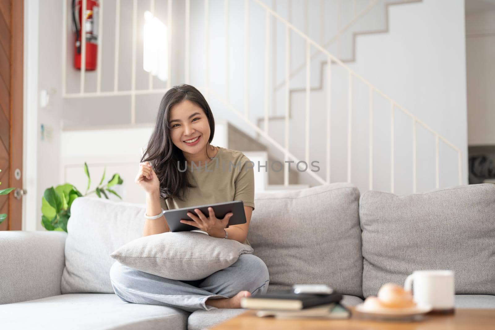 Beautiful attractive young Asian woman sitting on gray sofa and using digital tablet at living room in the morning