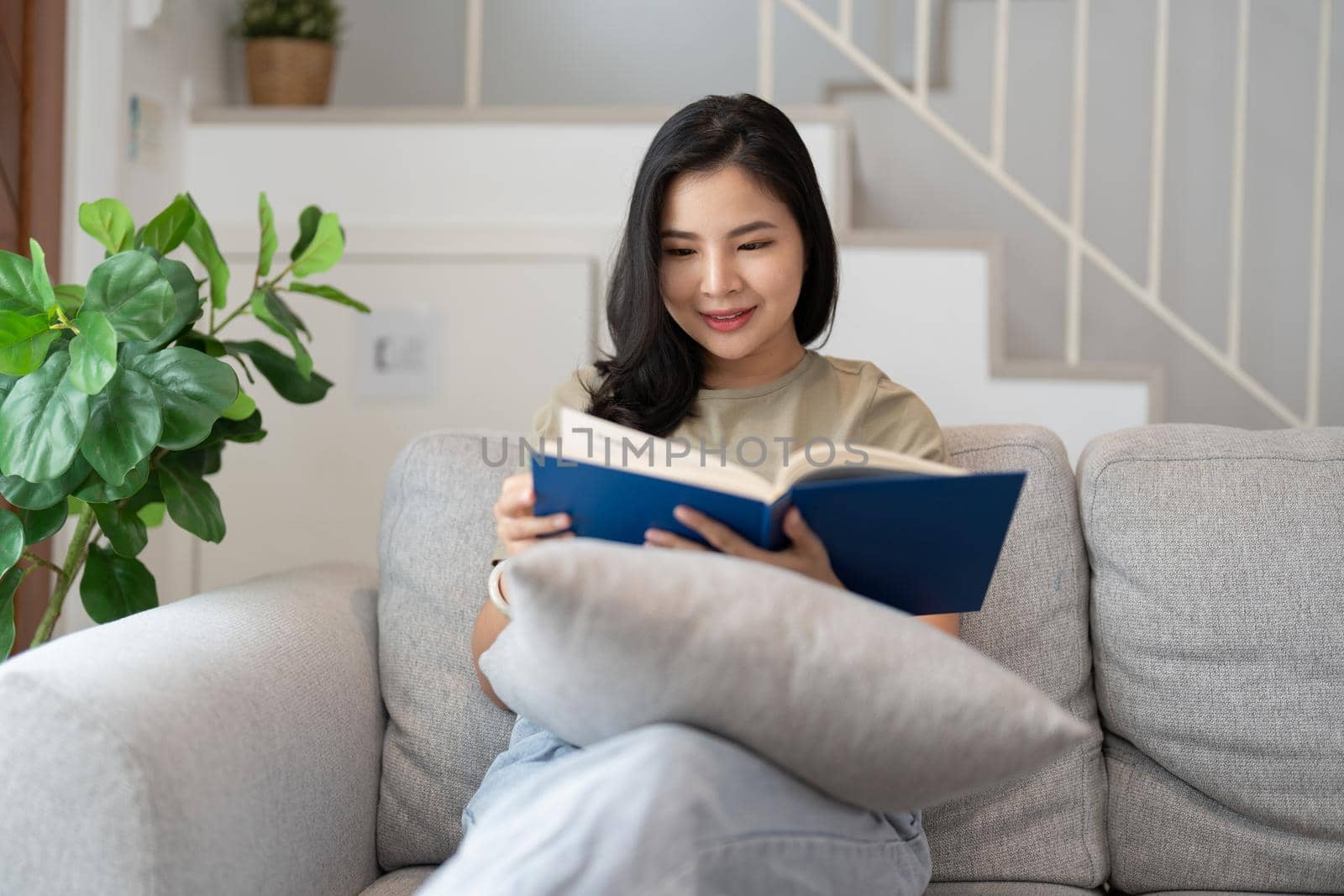 happy asian young woman sitting on sofa and reading book at home -people and leisure concept by nateemee