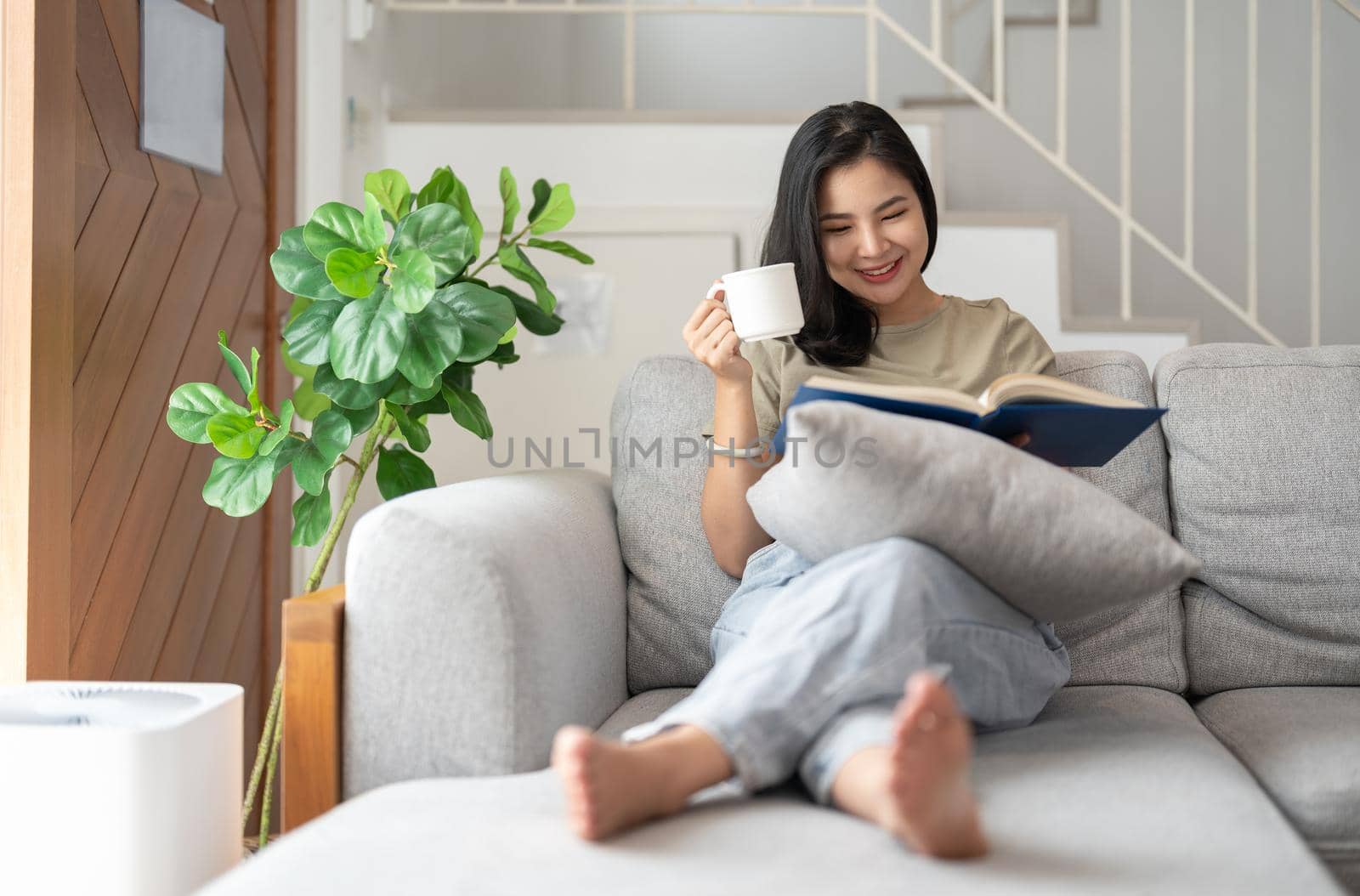 happy asian young woman sitting on sofa and reading book at home -people and leisure concept by nateemee