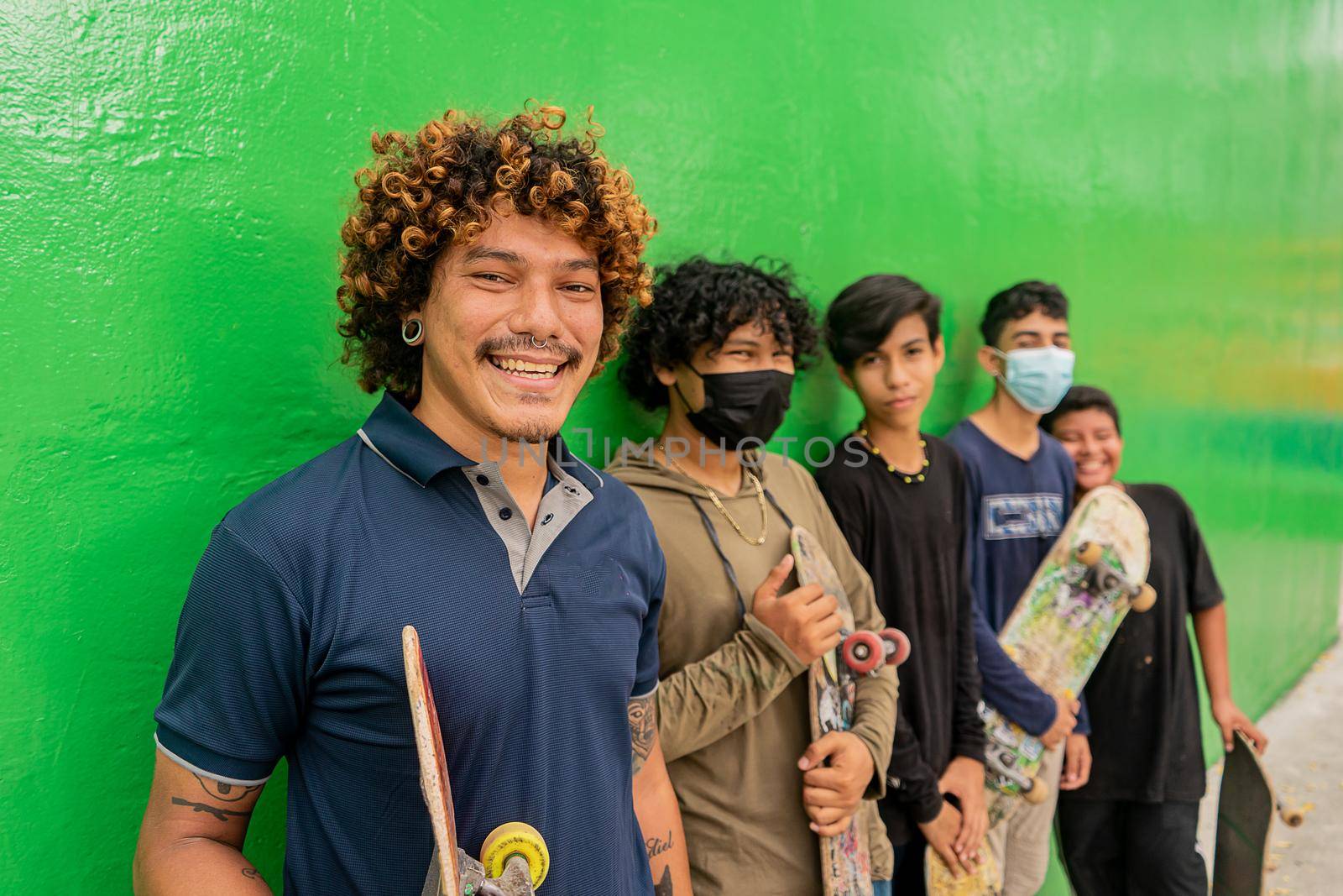 Group of skater teenagers and children smiling and holding their boards in front of a green wall