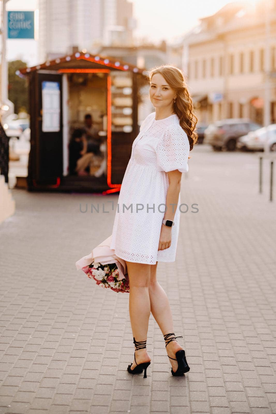 A happy woman in a white dress at sunset with a bouquet of flowers in the city.