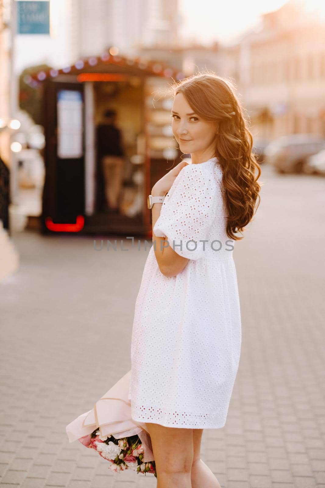 A happy woman in a white dress at sunset with a bouquet of flowers in the city.